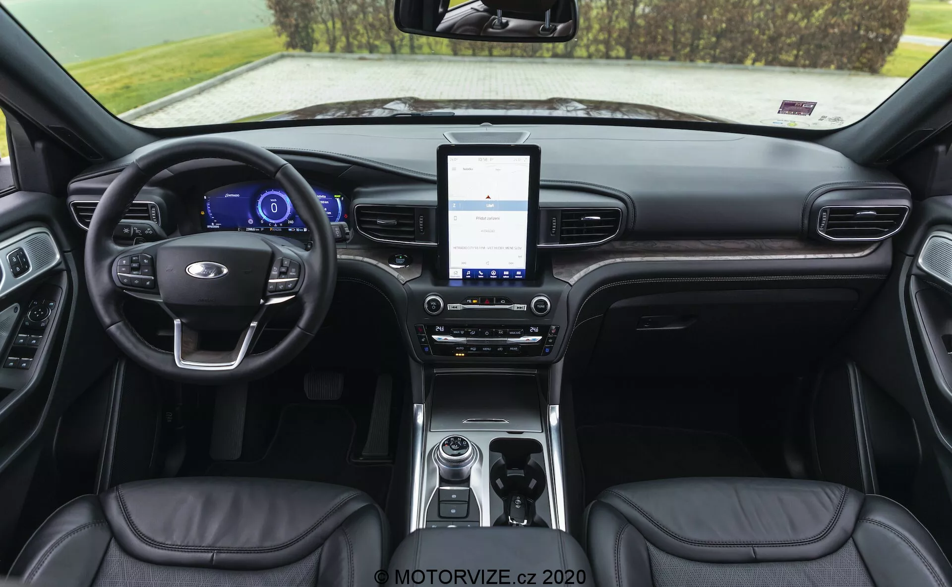 Interiér kabíny vozidla Ford Explorer 2019 zobrazujúci oblasť prístrojovej dosky z pohľadu smerom dozadu. Pohľad zahŕňa kožený volant s integrovanými ovládacími prvkami, digitálny prístrojový panel, veľký dotykový infotainment systém, dvojzónové klimatizačné ovládanie, otočný volič prevodovky, úložný priestor s konektivitnými možnosťami, kožené sedadlá s viditeľným prešitím a čiastočne viditeľné panely dverí s ovládacími prvkami.
