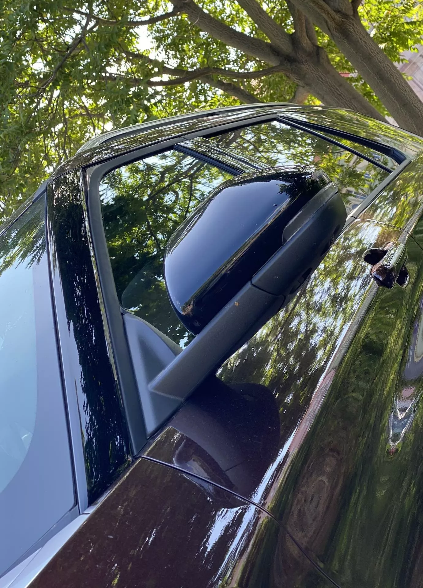 Detailný, šikmý pohľad na dvere a spätné zrkadlo na strane vodiča u vozidla Ford Explorer 2019. Fotografia vyhotovená mierne nad úrovňou rukoväte dverí odhaľuje lesklý tmavý exteriér, pravdepodobne čierny alebo tmavomodrý, s odrazmi naznačujúcimi čistú, dobre udržiavanú karosériu. Sklopené zrkadlo v zodpovedajúcej farbe naznačuje vyšší model. Viditeľné čiastočne stiahnuté okno a moderná rukoväť, pravdepodobne s dotykovým alebo bezklúčovým vstupom. Odrazy listov a vetiev naznačujú parkovanie vonku.