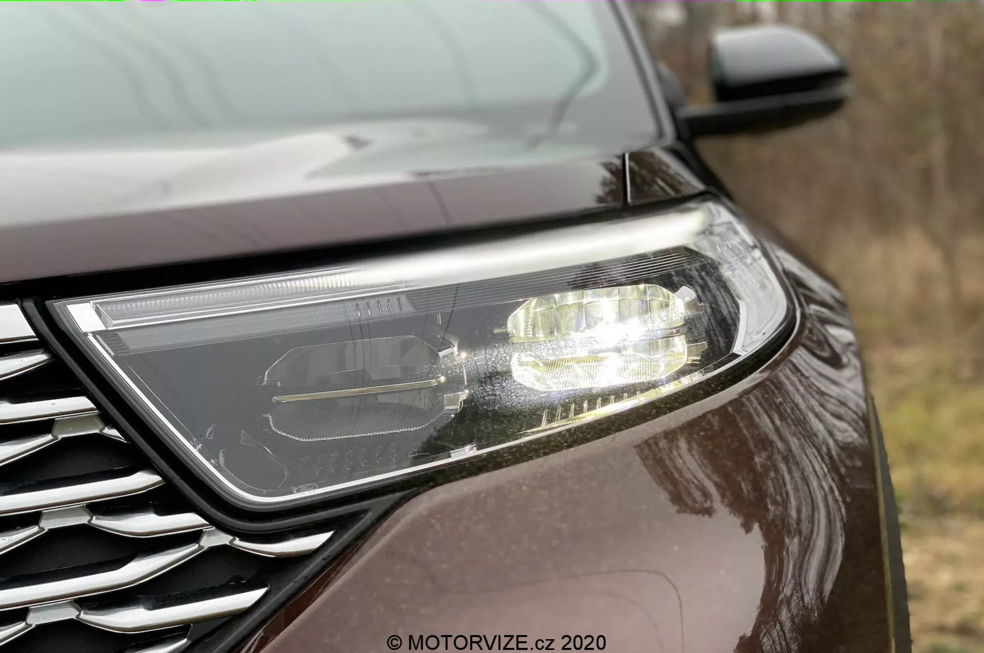 Detailný bočný pohľad na prednú časť Fordu Explorer 2019. Výrazný moderný svetlomet naznačujúci LED alebo HID technológiu s aktívnymi denné svetlá. Čiastočne viditeľná mriežka s kovovým odznakom, umiestnená v nízkom uhle. Auto má tmavý, reflektívny náter, čo naznačuje metalický alebo perleťový povrch, v exteriéri.