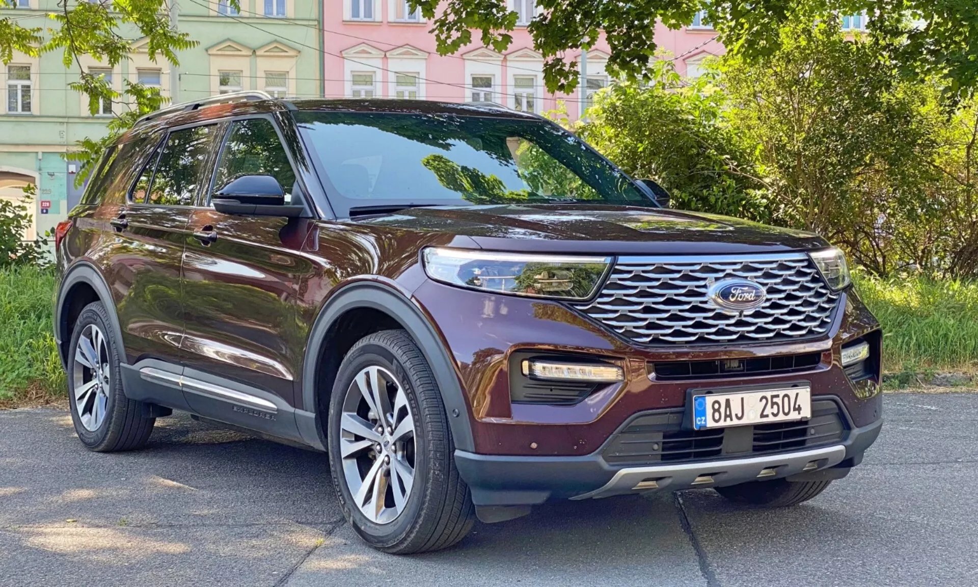 Trojštvrťový predný pohľad na moderné hnedé SUV Ford Explorer 2019, ktorý zobrazuje jeho bočný profil a prednú časť. Vozidlo je zaparkované na mestskej ceste s budovami a stromami v pozadí. Viditeľné kľúčové črty zahŕňajú výraznú prednú mriežku s horizontálnymi lamelami a centrálnym emblémom, moderné LED denné svetlá, hmlové svetlá integrované v dolnom nárazníku, veľké viacramenné zliatinové kolesá, bočné zrkadlá s integrovanými smerovými svetlami, tmavé okná, strešné lišty, tvarované línie karosérie a zvýšenú svetlú výšku.