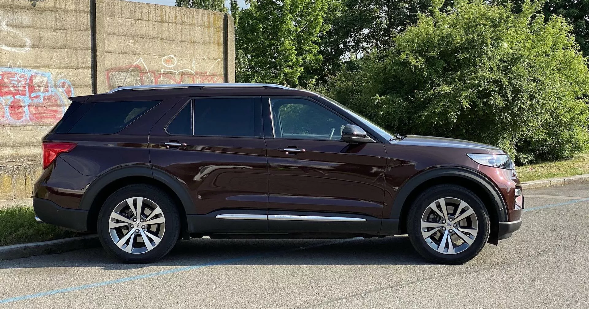 Moderné SUV Ford Explorer 2019 z bočného pohľadu, zaparkované na dláždenej ploche vedľa obrubníka s graffiti pokrytou stenou v pozadí, čo naznačuje mestské prostredie. Vlastnosti zahŕňajú elegantné svetlomety, výraznú charakteristickú líniu, bočné zrkadlá s indikátormi smerových svetiel, päťramenné kolesá z ľahkých zliatin, chrómové prvky okolo okien, strešné lišty, tónované zadné okná, lakované kľučky dverí a dostatočnú svetlú výšku. Fotografia za denného svetla zobrazuje celý profil vozidla.