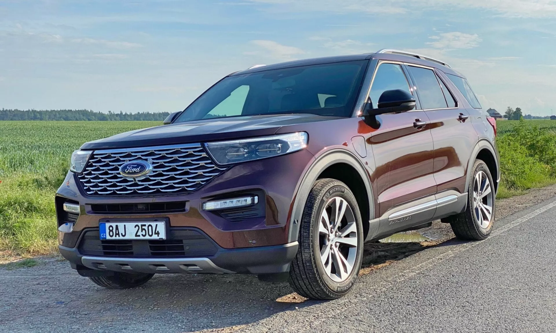Pohľad na tri štvrtiny zpredu na SUV Ford Explorer 2019. Obrázok zobrazuje prednú mriežku vozidla so sieťovým vzorom, elegantné, predĺžené svetlomety s viditeľnými LED dennými svetlami, tvarovanú kapotu so zdvihnutými kontúrami, zliatinové kolesá s viacramenným dizajnom a farebne zladené bočné zrkadlá s integrovanými smerovými svetlami. Okna majú chrómovaný alebo strieborný lem. Vozidlo je kovovej bordovej farby a nachádza sa na asfaltovej ceste v trávnatej, potenciálne vidieckej oblasti.