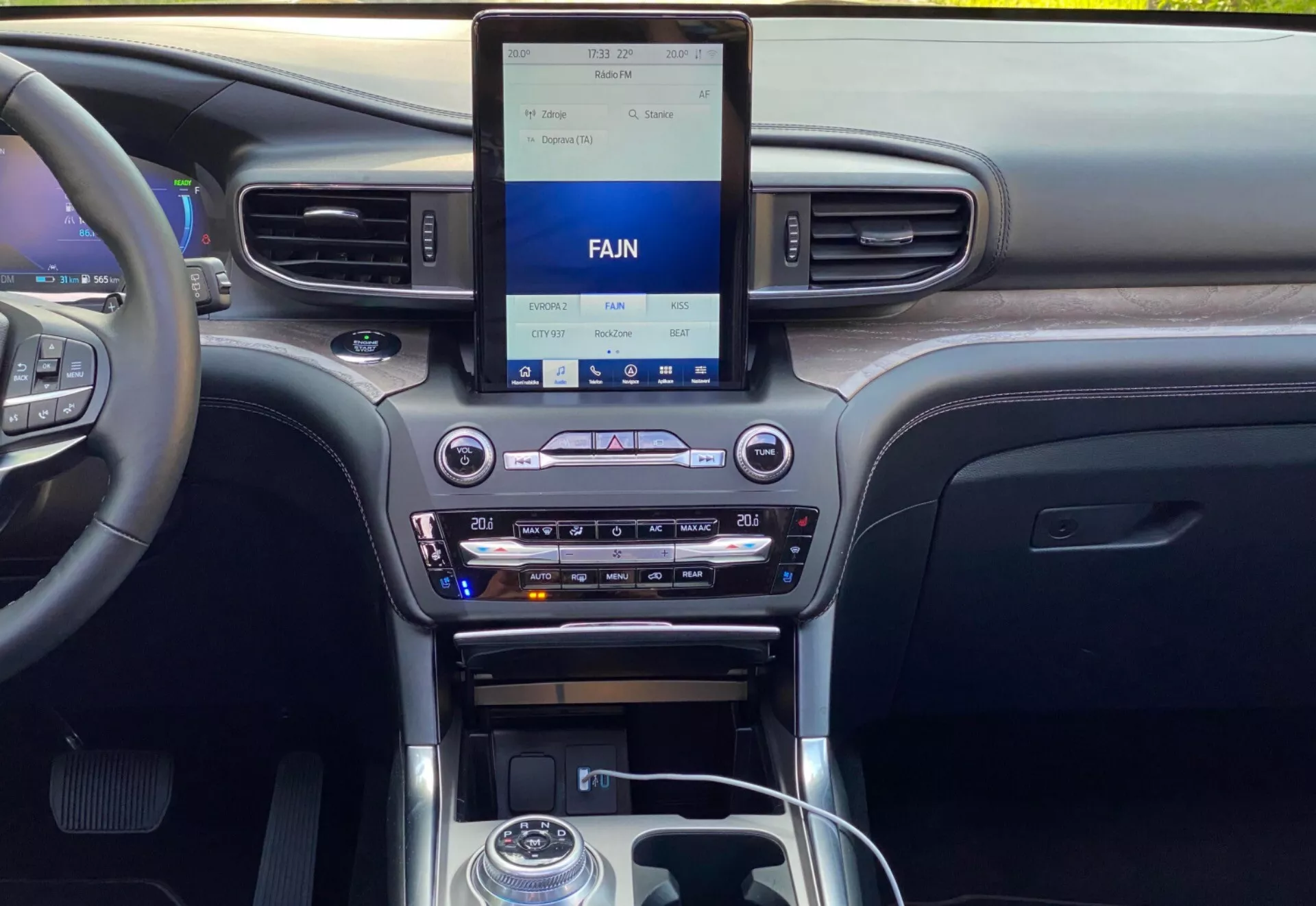 Interiér Ford Explorer 2019, zobrazujúci modernú centrálnu palubnú dosku z pohľadu predného alebo zadného sedadla. Palubná doska obsahuje vertikálny infotainment dotykový displej s ovládaním klimatizácie nižšie, vrátane nastavení duálnej klimatizácie, slotu na CD a možnosti pripojenia médií. Centrálna konzola zahŕňa radiacu páku s kovovým akcentom, ovládacie tlačidlá, otočný volič a držiaky nápojov. Materiály naznačujú kombináciu dreva, kovových úprav a čierneho plastu, zdôrazňujúc technológiu a kvalitný dizajn interiéru vozidla.