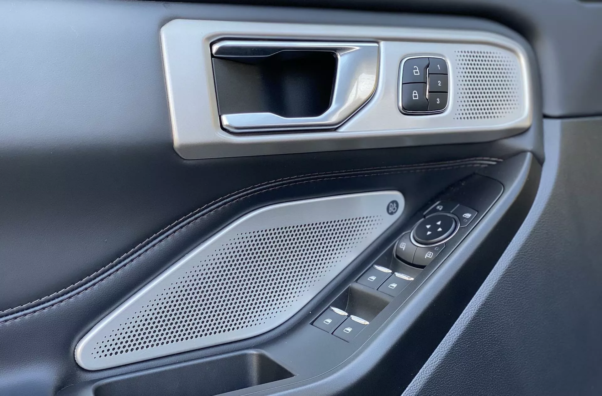 Obraz zachytáva interiérový panel dverí na strane vodiča vozidla Ford Explorer 2019. Pohľad z mierne zvýšenej a mierne naklonenej perspektívy ukazuje kovovo vyzerajúcu kľučku dverí osadenú v ráme, vedľa ktorej je tlačidlo zámku/odomknutia vložené do jemne textúrovaného panela. Nižšie sa nachádza oválna mriežka reproduktora s okrúhlymi otvormi a niekoľko ovládacích tlačidiel a prepínačov pre bočné zrkadlá a okná, čo naznačuje elektrické okná pre viacero dverí. Tmavá farebná paleta s kontrastnými kovovými prvkami a viditeľným šitím na pravdepodobne koženej opierke ruky zvýrazňuje pocit luxusu. Plytká hĺbka ostrosti zvýrazňuje ovládacie prvky, zdôrazňujúc dizajn a umiestnenie.