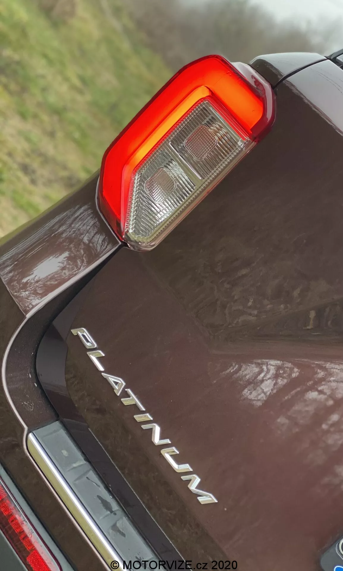 Detailný záber z nízkeho uhla na zadnú časť Ford Explorer 2019 PLATINUM s dôrazom na vertikálny LED zadný svetlomet, s čírou a červenou sekciou pre cúvacie a brzdové svetlá, chrómovým nápisom 'PLATINUM', odrazivou tmavou farbou s kvapkami vody a chrómovú lištu pod zadným nárazníkom.