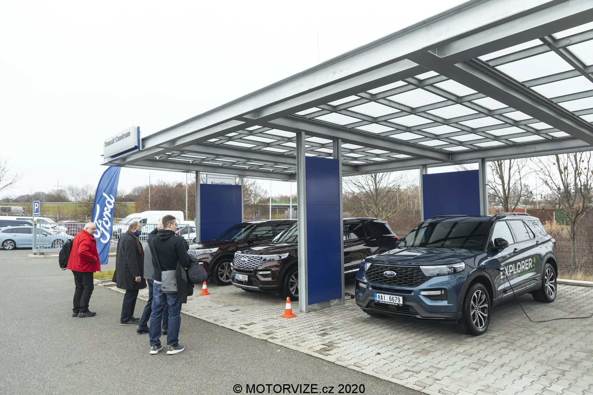 Obrázok zachytáva vonkajší autosalón alebo výstavnú udalosť pod kovovou, polopriehľadnou strechou so žiarivým, zamračeným denným svetlom. Tri SUV, vrátane moderného Ford Explorer 2019 v popredí, sú vystavené s viditeľnými prvkami ako predná maska, svetlomety a bočné zrkadlá. Ľudia a bannery naznačujú oficiálnu propagačnú akciu, logo Ford je rozpoznateľné v pozadí.