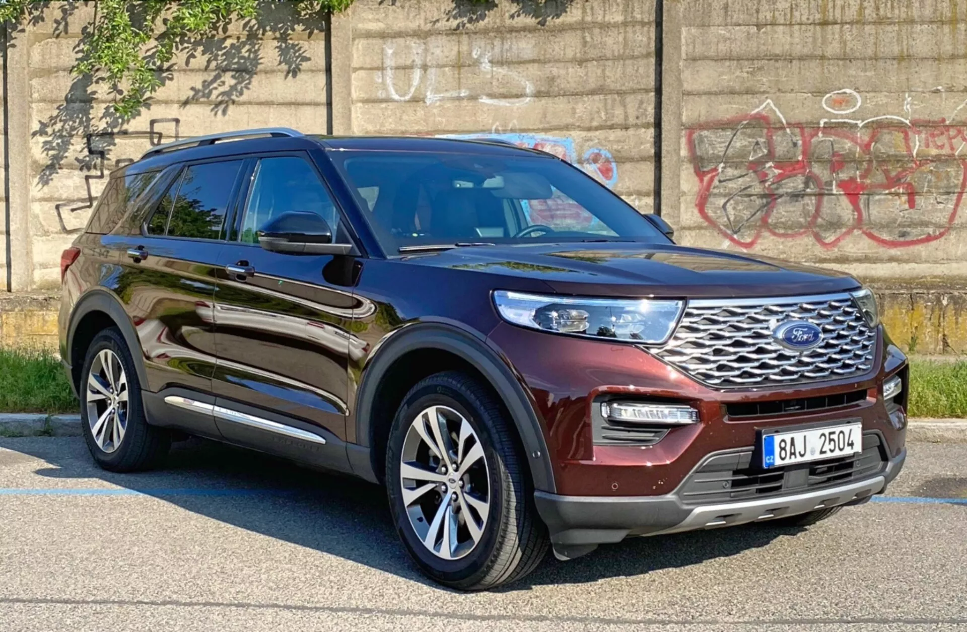 Obrázok SUV, konkrétne Ford Explorer 2019, nasnímaný z trojštvrťového predného uhlu od strany vodiča, ukazujúci lesklú kovovo-hnedú alebo burgundskú farbu. Obrázok zvýrazňuje prednú mriežku s logom Ford, elegantné LED alebo HID svetlomety, predný nárazník a profil vodičovej strany vrátane dverí, blatníka a viacramenných zliatinových kolies. Vozidlo je zaparkované v mestskej oblasti s graffiti na betónovej stene v pozadí, zdôrazňujúc jeho robustný a moderný dizajn, s možnosťou pokročilých asistenčných systémov pre vodiča, ako je adaptívny tempomat a asistent udržiavania v jazdnom pruhu.