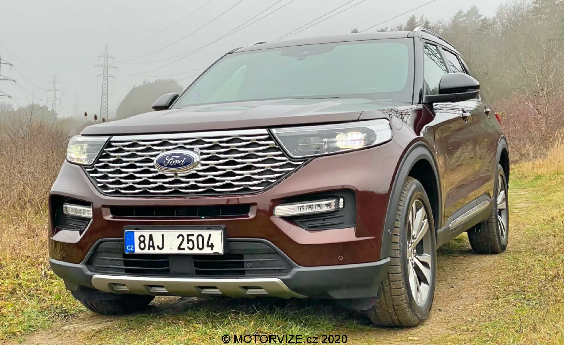 2019 Ford Explorer auto fotografované z nízkeho predného uhla tri štvrtiny, zobrazujúce mriežku s logom Ford, svetlomety, hmlové svetlá, kapotu, bočné okná a časť zadnej časti. Zložitý design mriežky, elegantné LED svetlomety, dvojfarebný nárazník a bezkľúčový vstupný systém zdôrazňujú moderné a technologicky vyspelé funkcie. Umiestnené mierne off-road s pravými kolesami na tráve a ľavými kolesami na ceste, zvýrazňujúcimi ľahké off-road schopnosti. Viditeľné viacramenné hliníkové kolesá, indikujúce celoročné pneumatiky.