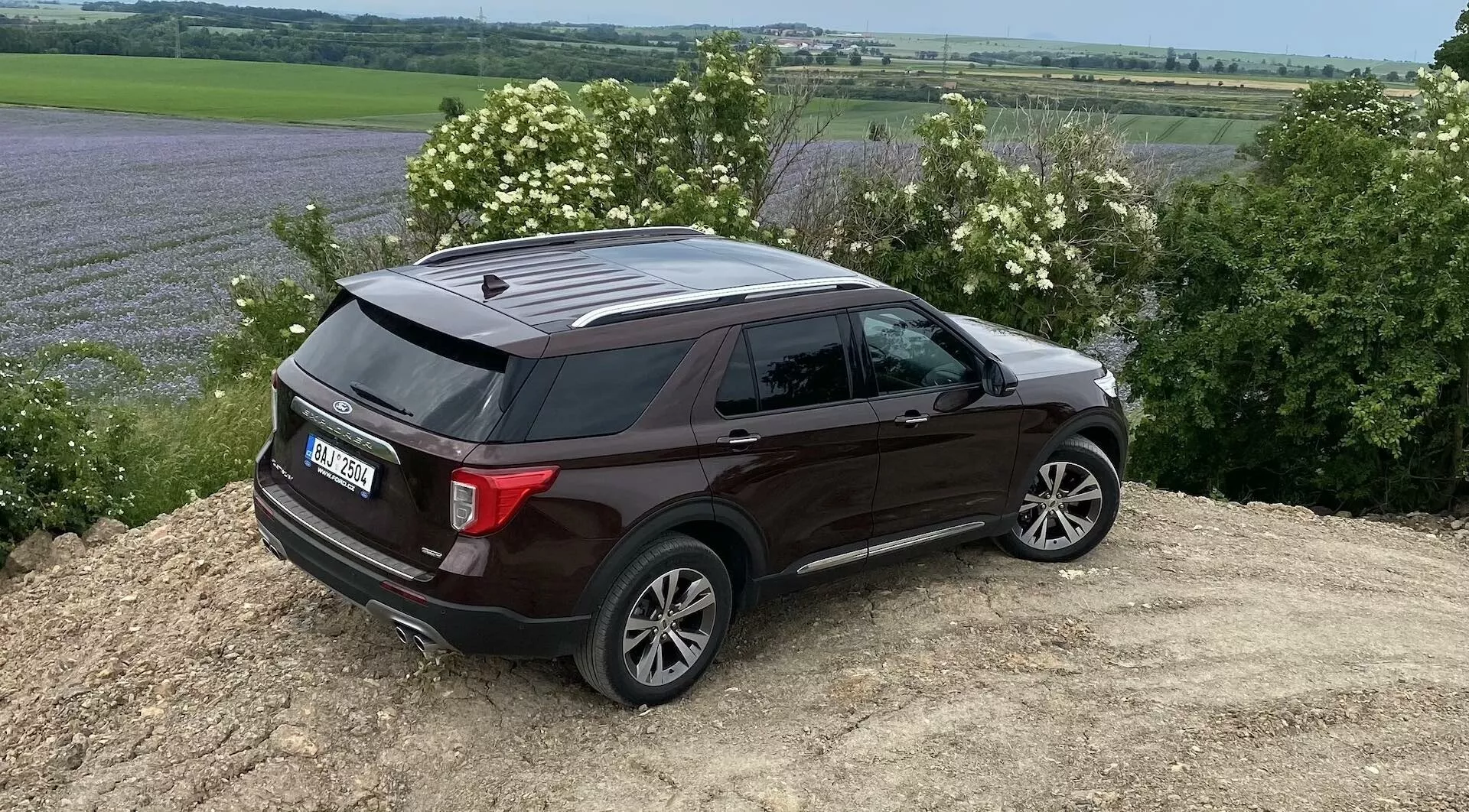 Ford Explorer 2019 v tmavej farbe zobrazený zo zadného trojštvrťového pohľadu, zaparkovaný na naklonenej hromade hliny. Kľúčové vlastnosti zahŕňajú viaclúčové zliatinové kolesá, tónované zadné okná s chrómovými lištami, strešné lyžiny, moderné zadné svetlá možne s LED technológiou, žraločiu anténu na streche a zadný spojler. Vidiecke prostredie s otvorenými poliami a stromami zdôrazňuje jeho schopnosti v teréne. Prirodzené svetlo a zamračená obloha vytvárajú pod autom mäkké tiene.