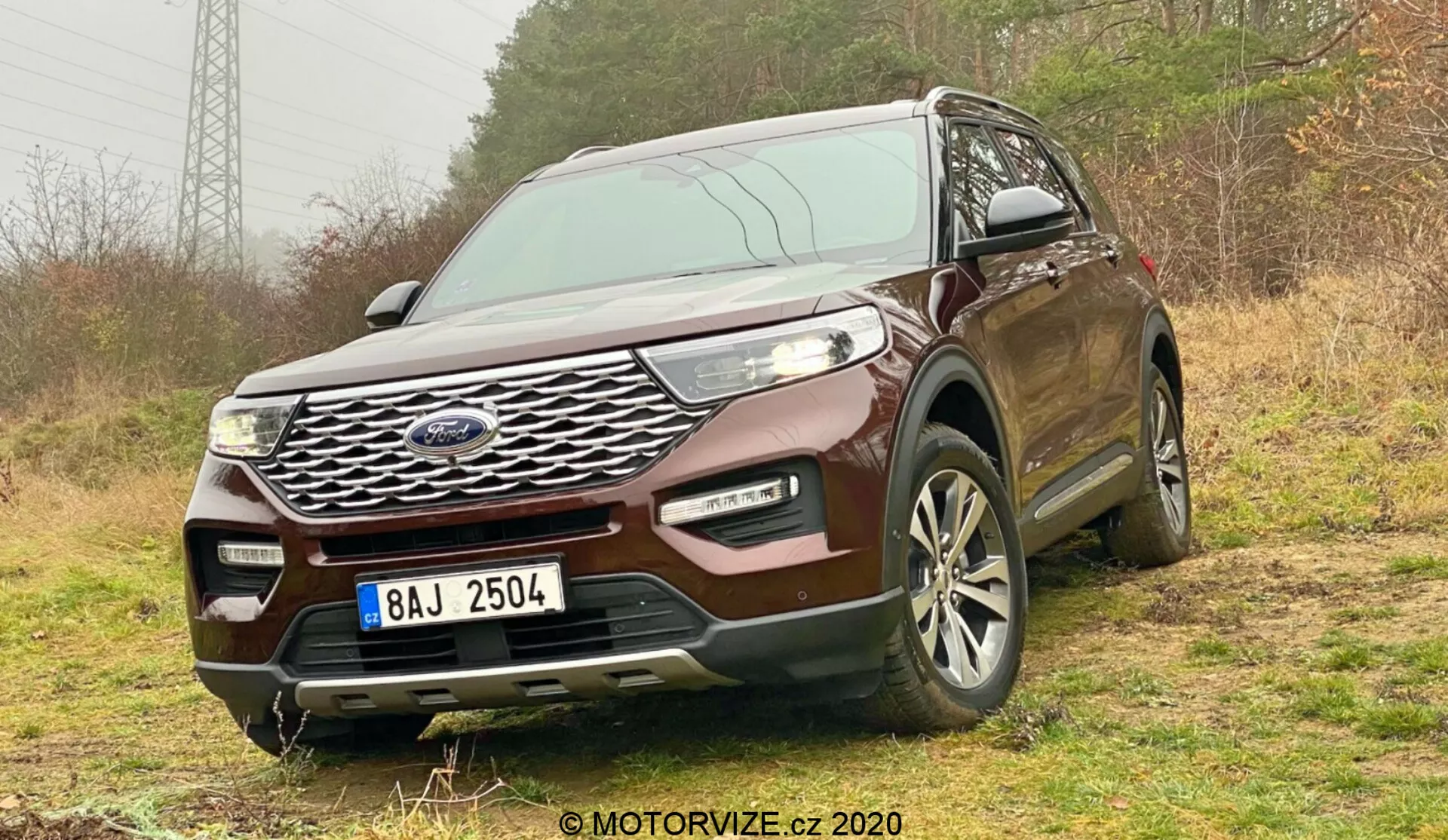 Predný trojštvrťový pohľad na SUV Ford Explorer 2019 urobený z nízkeho uhla, čo zvýrazňuje jeho výraznú chrómovanú mriežku, elegantné LED svetlomety a viacramenné zliatinové kolesá. SUV je umiestnené na nespevnenej ploche s vegetáciou, čo naznačuje jeho spôsobilosť do terénu. Predný nárazník obsahuje hmlové svetlá a prívody vzduchu, čo naznačuje zvýšenú funkčnosť. Senzory alebo kamery môžu naznačovať pokročilé bezpečnostné funkcie. ŠPZ je začiernená a v pravom dolnom rohu je vodoznak 