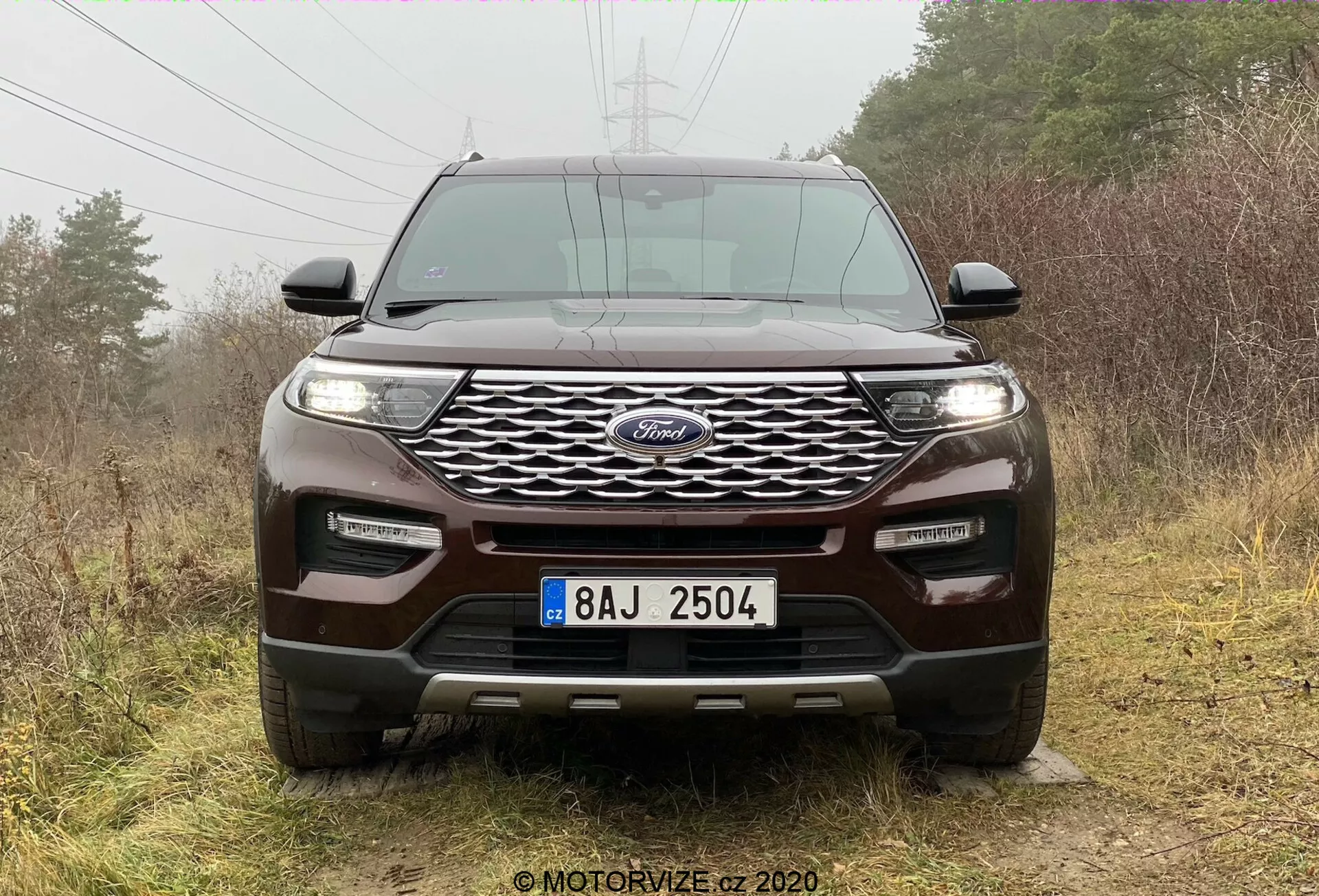 Čelný pohľad na SUV Ford Explorer 2019 s výrazným modrým oválnym znakom uprostred dynamickej mriežky, lemovanej elegantnými horizontálne orientovanými svetlometmi pravdepodobne s LED alebo HID žiarovkami. Integrované hmlové svetlá a parkovacie senzory zvyšujú technologické a bezpečnostné prvky predného nárazníka. Tvarovaná kapota so zvýšenými hrebeňmi zdôrazňuje športový dizajn, zatiaľ čo značná svetlá výška, ochranný plech a ťažné háky naznačujú schopnosti v teréne. SUV je zasadený do vonkajšieho, prírodného pozadia s mdlo rozptýleným svetlom, ktoré zvýrazňuje jeho dizajn.