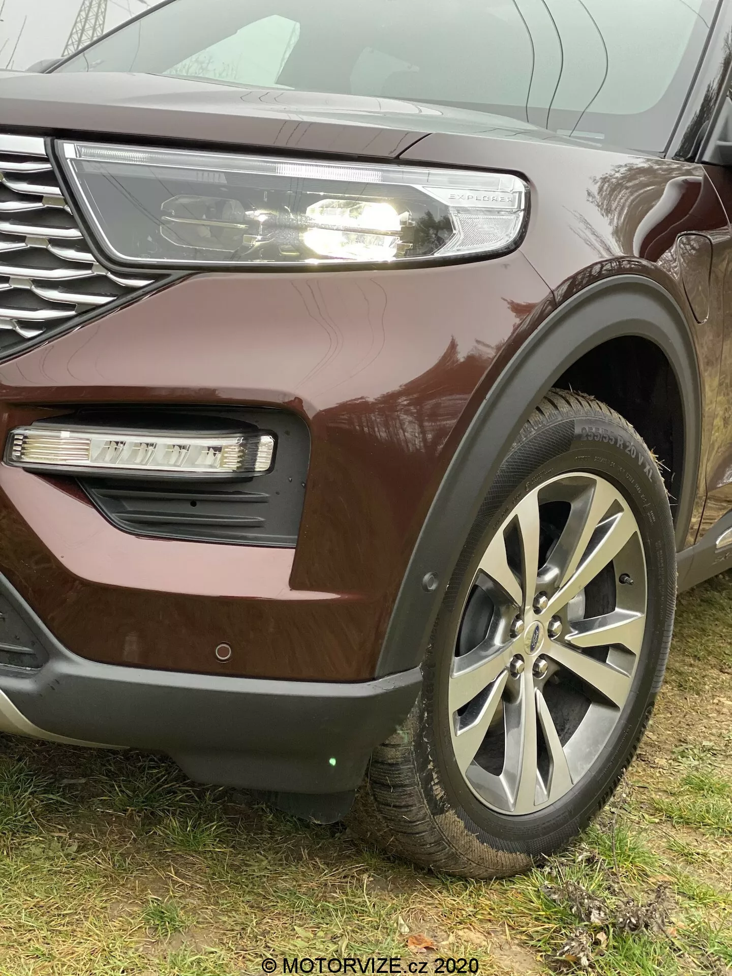 Detailný, šikmý pohľad na SUV Ford Explorer 2019 so zameraním na ľavý predný roh, zdôrazňujúci veľké zliatinové kolesá, masku chladiča s chrómovými akcentmi a čiastočne viditeľné LED svetlomety s dennými svetlami, kovový burgundský lak, hmlové svetlo v zapustenej oblasti nárazníka s odlišnou textúrou a možné sacie otvory alebo dekoratívne lišty. Logo je v strede viaclúčových kolies. Spodná časť nárazníka naznačuje prítomnosť parkovacieho senzora alebo adaptívneho tempomatu. Obrázok bol urobený z nízkeho uhla, aby zdôraznil veľkosť a robustnosť vozidla, s vodoznakom 'MOTORVIZE.cz 2020' viditeľným v pravom dolnom rohu.