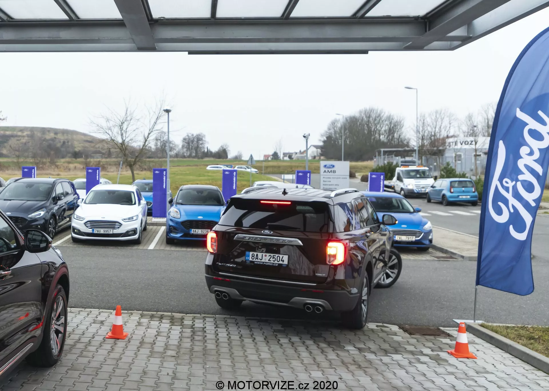 Čierne SUV Ford Explorer 2019, umiestnené v strede, buď cúva, alebo odchádza z EV nabíjacieho miesta v nabíjacej stanici. Zadné svetlá sú zapnuté, čo môže naznačovať cúvanie alebo brzdenie, s viditeľnou registračnou značkou, dvojitým športovým výfukom, zadným stieračom a vysoko umiestneným zadným brzdovým svetlom. Okolie ukazuje niekoľko elektrických áut pri modrých nabíjacích stĺpoch a oranžové dopravné kužele blízko SUV. Vlajka s logom Fordu naznačuje prítomnosť značky. Scéna je zachytená v zamračený deň s rozptýleným prírodným svetlom, zvýrazňujúcim automobilovú technológiu a udržateľnú dopravnú infraštruktúru.