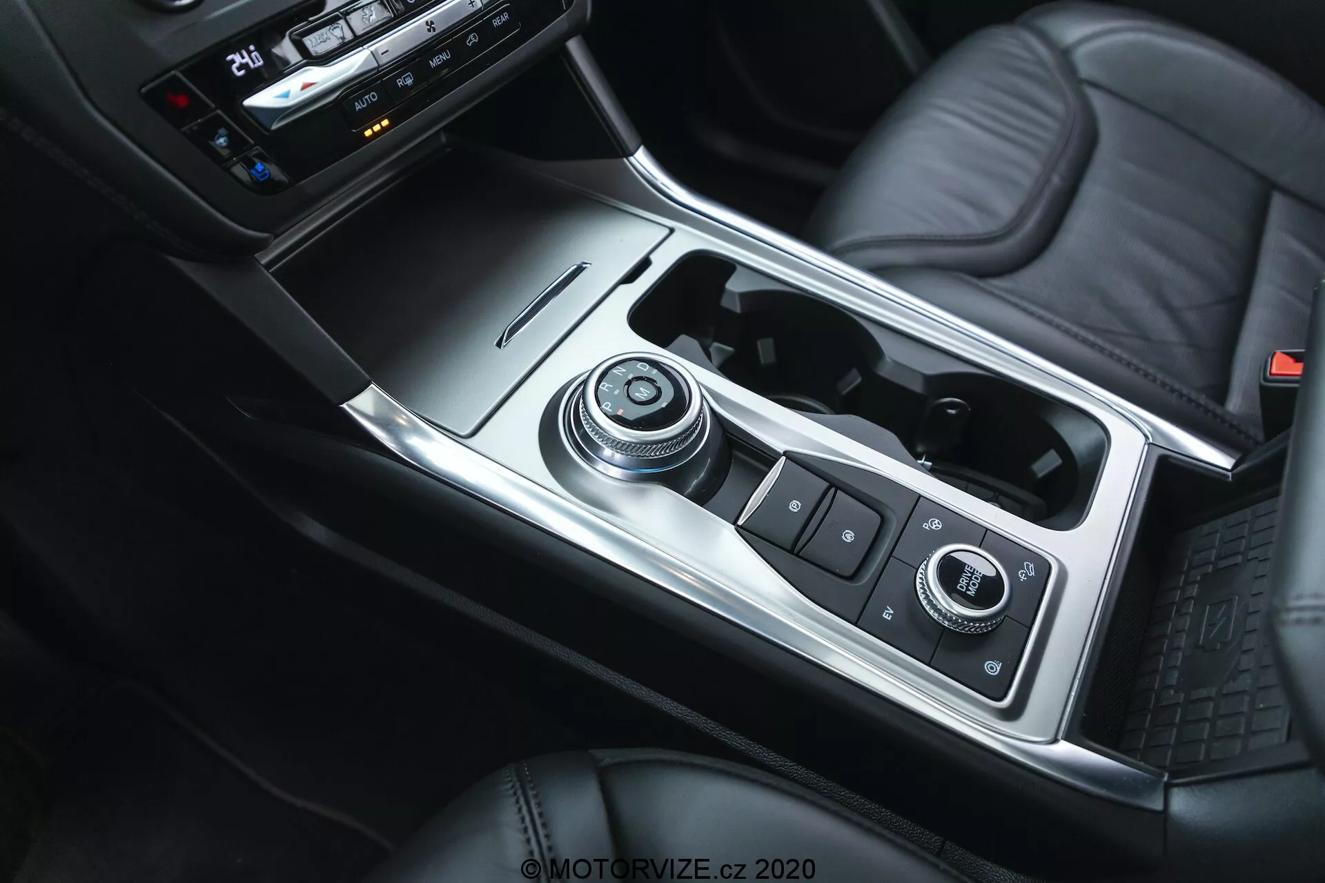 Interiérový záber na Ford Explorer 2019 zameraný na stredovú konzolu z pohľadu zhora, zobrazujúci radiacu páku, infotainment a ovládací systém vozidla, držiaky na nápoje, úložný priestor s USB portami, ovládacie prvky klimatizácie a stredovú lakťovú opierku čalúnenú kožou, s kombináciou kovových, kožených a plastových komponentov, zdôrazňujúce modernú estetiku a funkčnosť.