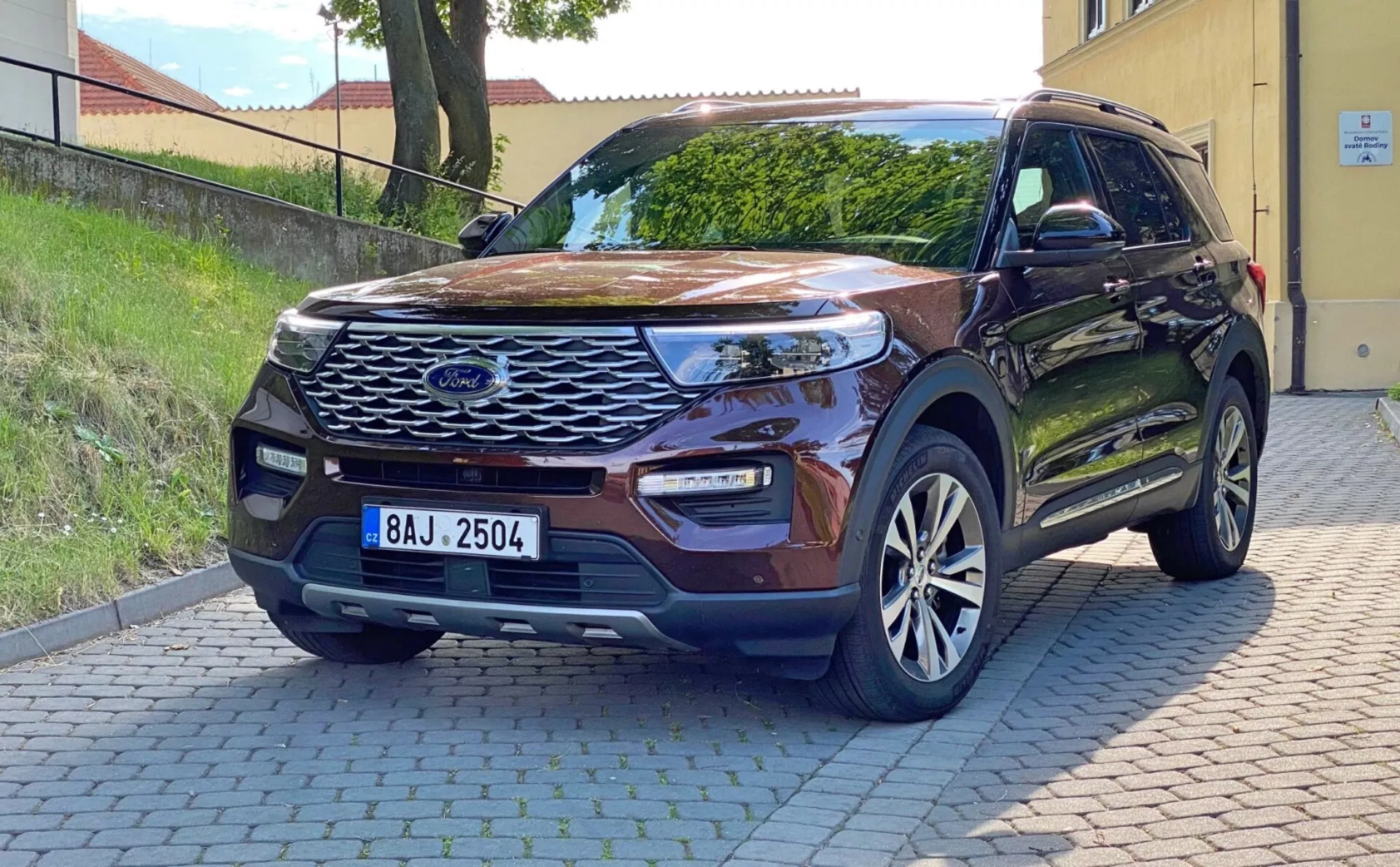 Obrázok moderného SUV Ford Explorer 2019 zaparkovaného na spevnenej ploche, zachytený z predného trojštvrtinového pohľadu. Ukazuje výraznú prednú mriežku s centrálnym znakom, elegantné LED svetlomety, integrované hmlové svetlá v nárazníku, parkovacie senzory, tvarovanú kapotu, hliníkové kolesá, farebne zladené spätné zrkadlá s blinkrami, bezkľúčový vstup a rezidenčné pozadie. Prirodzené denné svetlo zvýrazňuje jeho vonkajšie vlastnosti a technické pokroky.