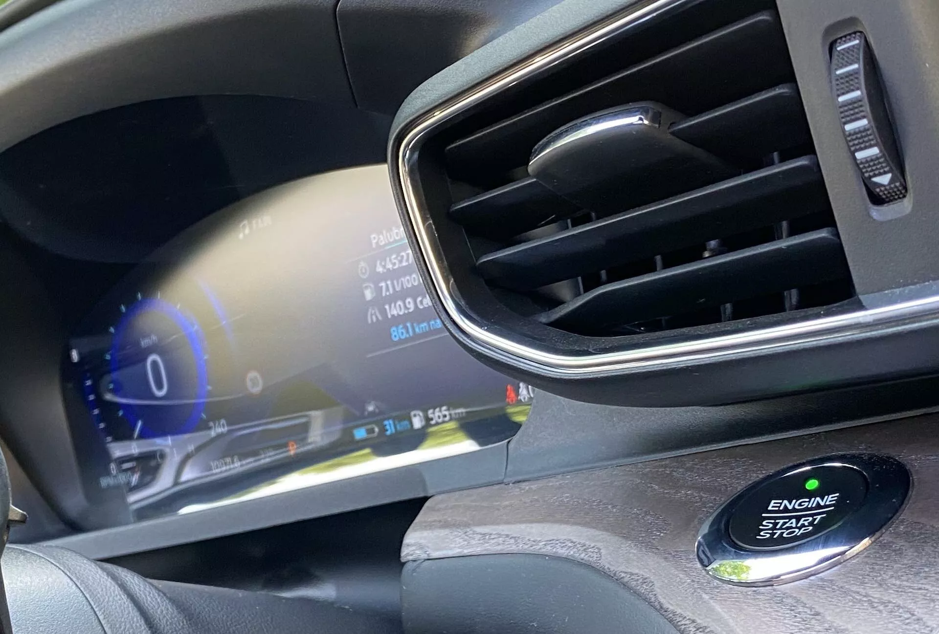 Detailný pohľad na interiér moderného automobilu, zameraný na prístrojovú dosku na strane vodiča. Obrázok zdôrazňuje digitálny prístrojový panel zobrazujúci stavy vozidla, ako je rýchlomer pri 0 km/h, ukazovateľ paliva a vonkajšiu teplotu 31 stupňov Celzia. Elegantný displej s vysokým rozlíšením ponúka živé farby a luxusný dizajn. Vpravo je vidieť vetrací otvor s chrómovaným akcentom a v pravom dolnom rohu mierne rozmazané tlačidlo ENGINE START STOP, ktoré naznačuje bezkľúčový štartovací systém.