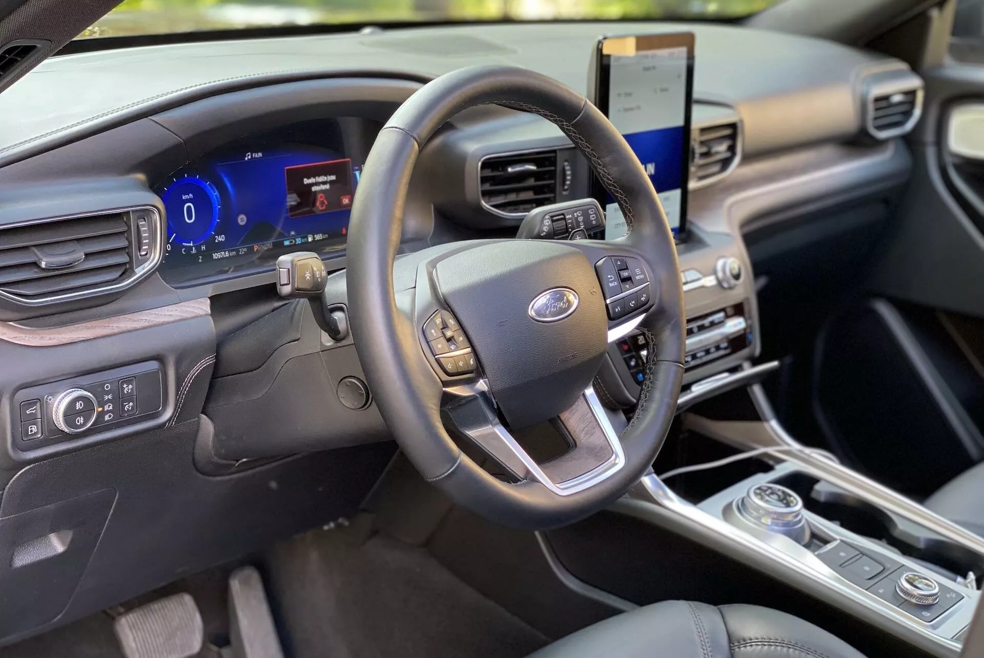 Interiér Fordu Explorer 2019, zameraný na oblasť vodiča z pohľadu predného spolujazdca. Volant s multifunkčnými ovládačmi pre audio, tempomat a hands-free komunikáciu je uprostred. Za ním sa nachádza digitálny prístrojový panel s možnosťou prispôsobenia a reálnymi údajmi o vozidle. Napravo vidíme vertikálne orientovaný dotykový displej s infotainment systémom, ktorý podporuje satelitnú navigáciu a integráciu chytrých telefónov. Dole sú ovládače klimatizácie s viacerými zónami a otočný volič prevodovky, čo naznačuje moderný dizajn. Kabína s čistou a modernou prístrojovou doskou, vyrobenou z prémiových materiálov, vyžaruje minimalistickú estetiku.