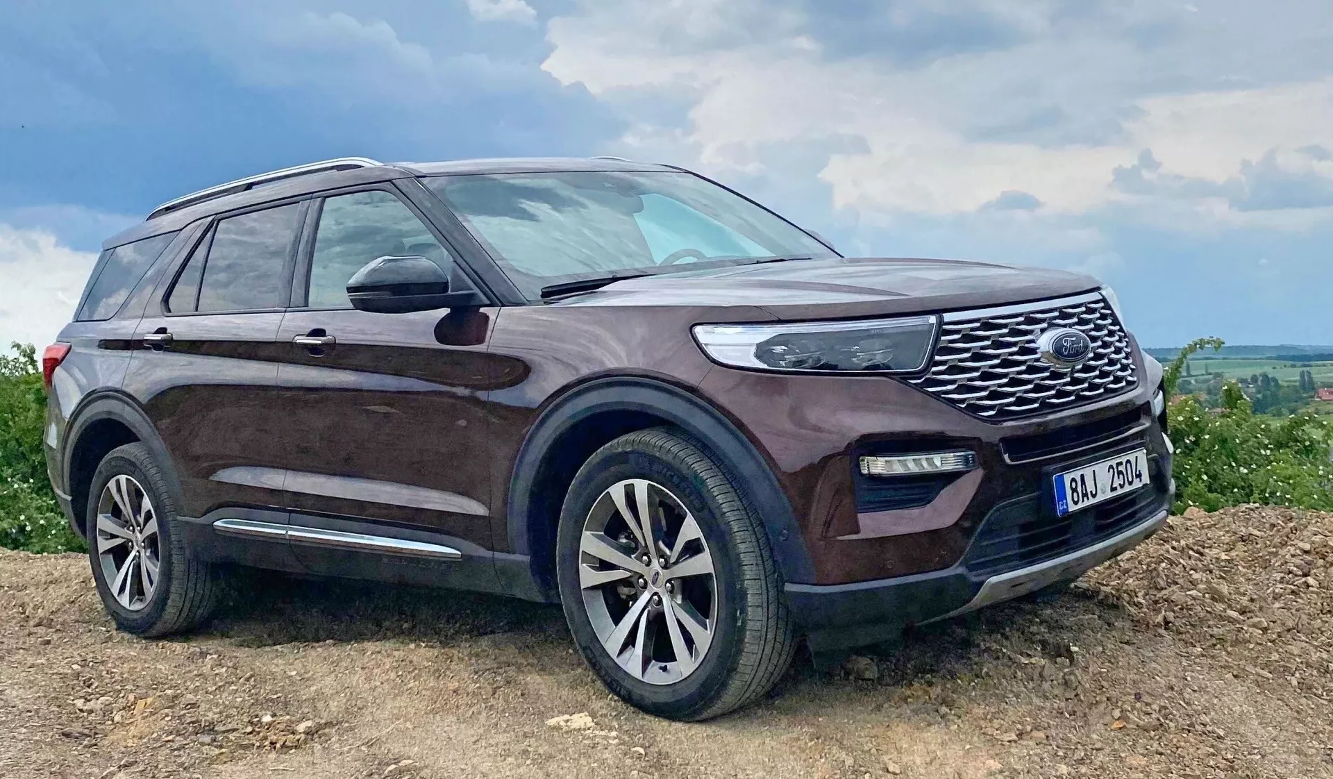 Obrázok SUV Ford Explorer 2019 zaparkovaného na naklonenej, možno nespevnenej ploche s tmavým odtieňom a zablatenými stranami, výraznou prednou mriežkou s mriežkovým dizajnom, LED svetlami, hmlovými svetlami, viaclúčovými zliatinovými kolesami, bočnými zrkadlami so zabudovanými smerovkami, strešnými lištami, bočným schodíkom, mierne tónovanými oknami a anténou na zadnej streche, zdôrazňujúc jeho odolné schopnosti a luxusné funkcie v vidieckom alebo off-road prostredí.