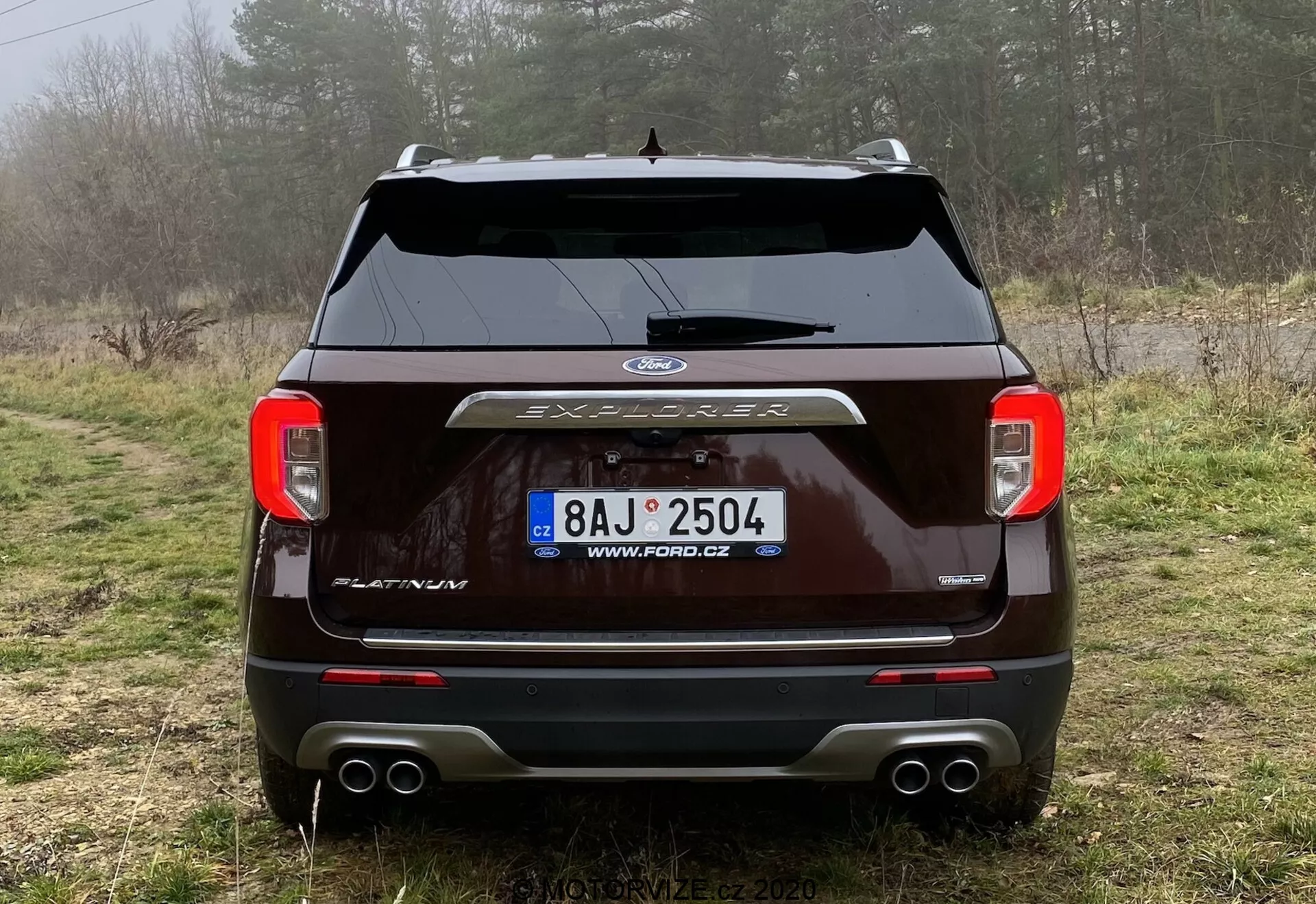 Zadný pohľad na tmavo sfarbený Ford Explorer 2019 zaparkovaný na poľnej ceste v vidieckom prostredí s vegetáciou a hmlou. Obrázok ukazuje jasne osvetlené zadné svetlá, centrálne umiestnené logo Ford, nápis 'EXPLORER', stierač zadného okna, parkovacie senzory, dvojitý výfuk a kryt ťažného zariadenia. Viditeľná je registračná značka s českou URL, čo zdôrazňuje dizajn exteriéru a technológie vozidla.