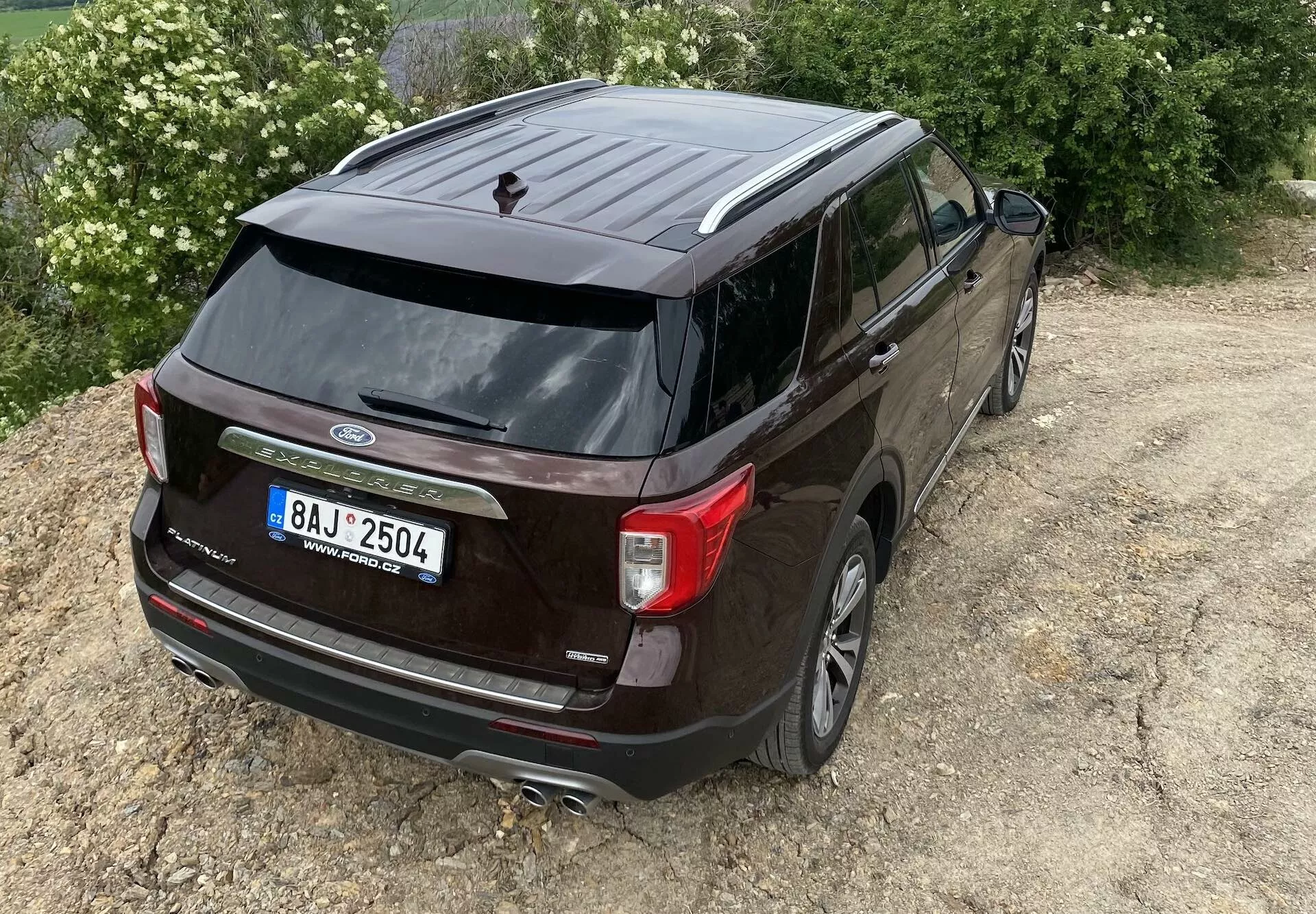 Vyvýšený pohľad na zadnú trojštvrť a tmavo sfarbený Ford Explorer 2019, zvýrazňujúci zadnú časť a bočný profil vozidla. Viditeľné funkcie zahŕňajú logo Ford, názov modelu 'Explorer', jedinečné zadné svetlá, stierač zadného okna, anténu v tvare žraločej plutvy, dvojitý výfukový systém, cúvaciu kameru, zliatinové kolesá, tónované okná a bočné zrkadlá s integrovanými smerovými svetlami. SUV je mierne mimo stred vpravo na neoznačenom teréne, čo naznačuje jeho schopnosť pre ľahké terénne použitie.