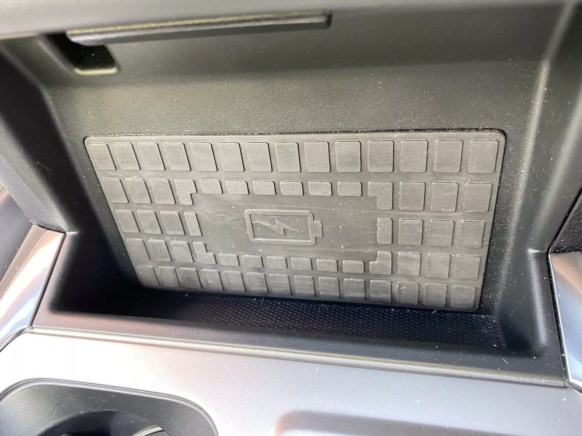 Detailný pohľad na priestor centrálnej konzoly Fordu Explorer 2019, zdôrazňujúci bezdrôtovú nabíjaciu podložku so zdvihnutou mriežkovou textúrou a protišmykovým povrchom. Viditeľné sú lesklé čierne plastové okraje, čo pridáva moderný automobilový dizajn. Slnečné svetlo naznačuje denné fotografovanie z predného sedadla, symbol nabíjania označuje pokročilú bezdrôtovú nabíjaciu technológiu.