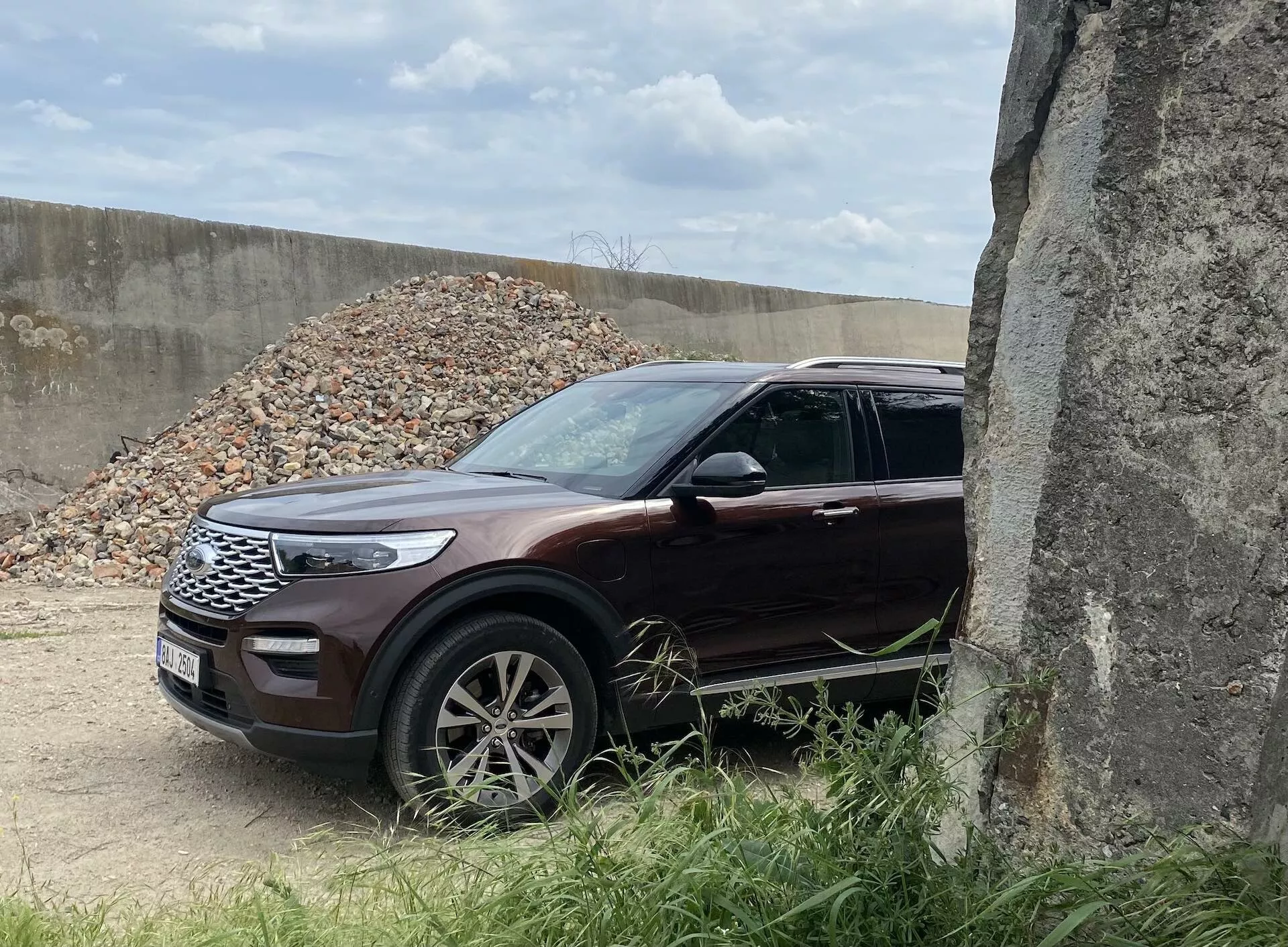 Obrázok zobrazuje SUV Ford Explorer z roku 2019 z trojštvrťového predného pohľadu, zvýrazňujúc prednú ľavú masku, elegantné LED svetlomety, kapotu, predné koleso a blatník. Vozidlo, lesklé hlboké bordovo červené alebo burgundské farby, stojí v exteriéri so štrkom v pozadí, čo naznačuje stavenisko. Viacramenné zliatinové kolesá, integrované smerové svetlá v zrkadlách a chrómové ozdoby pridávajú prémiový vzhľad. Čiastočne zakryté betónovou konštrukciou vpravo, toto SUV má moderný dizajn a bezpečnostné prvky, čo ho robí vhodným pre kategórie ako SUV, moderný dizajn áut, technológia vozidiel a terénne vozidlá.