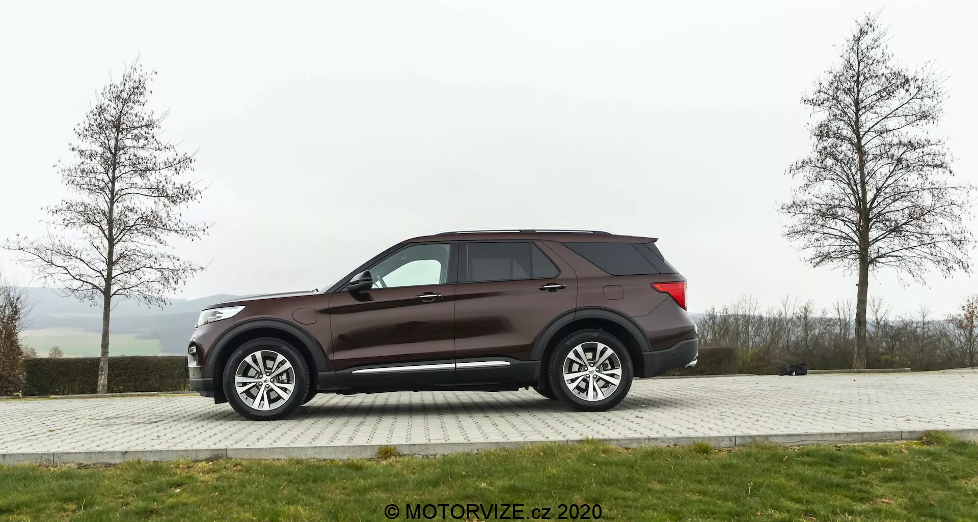 Bočný pohľad na SUV Ford Explorer z roku 2019, zaparkované na dláždenej ploche s trávnikom v pozadí za oblačného dňa. Vozidlo má veľké viacramenné zliatinové kolesá, tónované okná a tmavohnedú alebo bordovú farbu. Dizajnové prvky zahŕňajú bočné zrkadlá pravdepodobne s elektrickým nastavením a integráciou smeroviek, aerodynamickú líniu strechy a viditeľnú zadnú anténu.