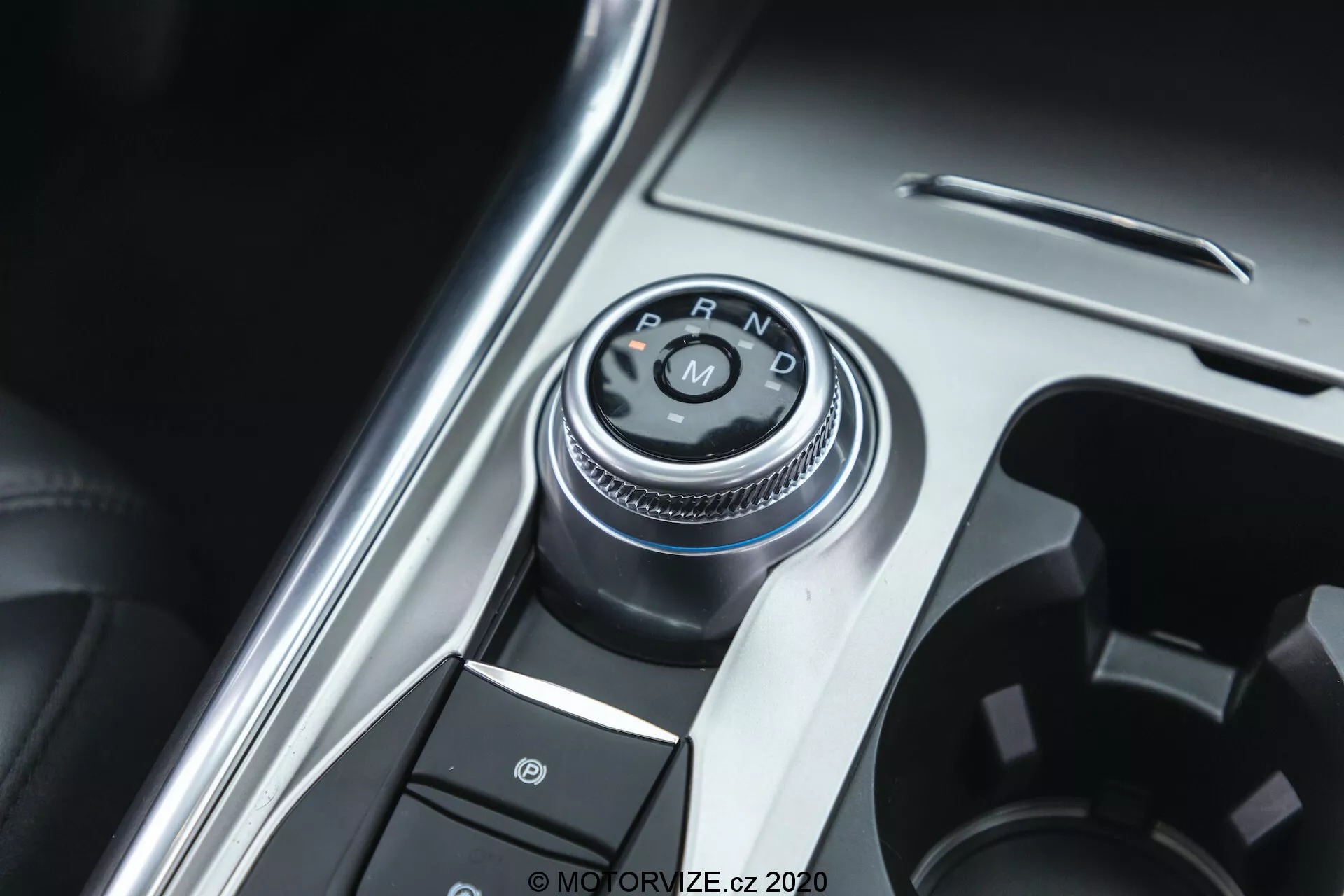 Blízky pohľad na selektor prevodovky a časť stredovej konzoly v interiéri Ford Explorer 2019. Hlavným bodom je rotačný selektor s vygravírovanými označeniami 'P, R, N, D', naznačujúcimi automatickú prevodovku. Fotografia je urobená z mierne nevycentrovaného, horného pohľadu. Okolo selektora prevodovky sú tlačidlá, vrátane jedného so symbolom parkovacej brzdy, čo naznačuje systém elektronickej parkovacej brzdy. Konzola kombinujúca kovové a plastové povrchy zdôrazňuje prémiový interiér vozidla. Dobré osvetlenie zvýrazňuje kovové prvky s malou hĺbkou ostrosti, ktorá sa sústreďuje na selektor prevodovky.