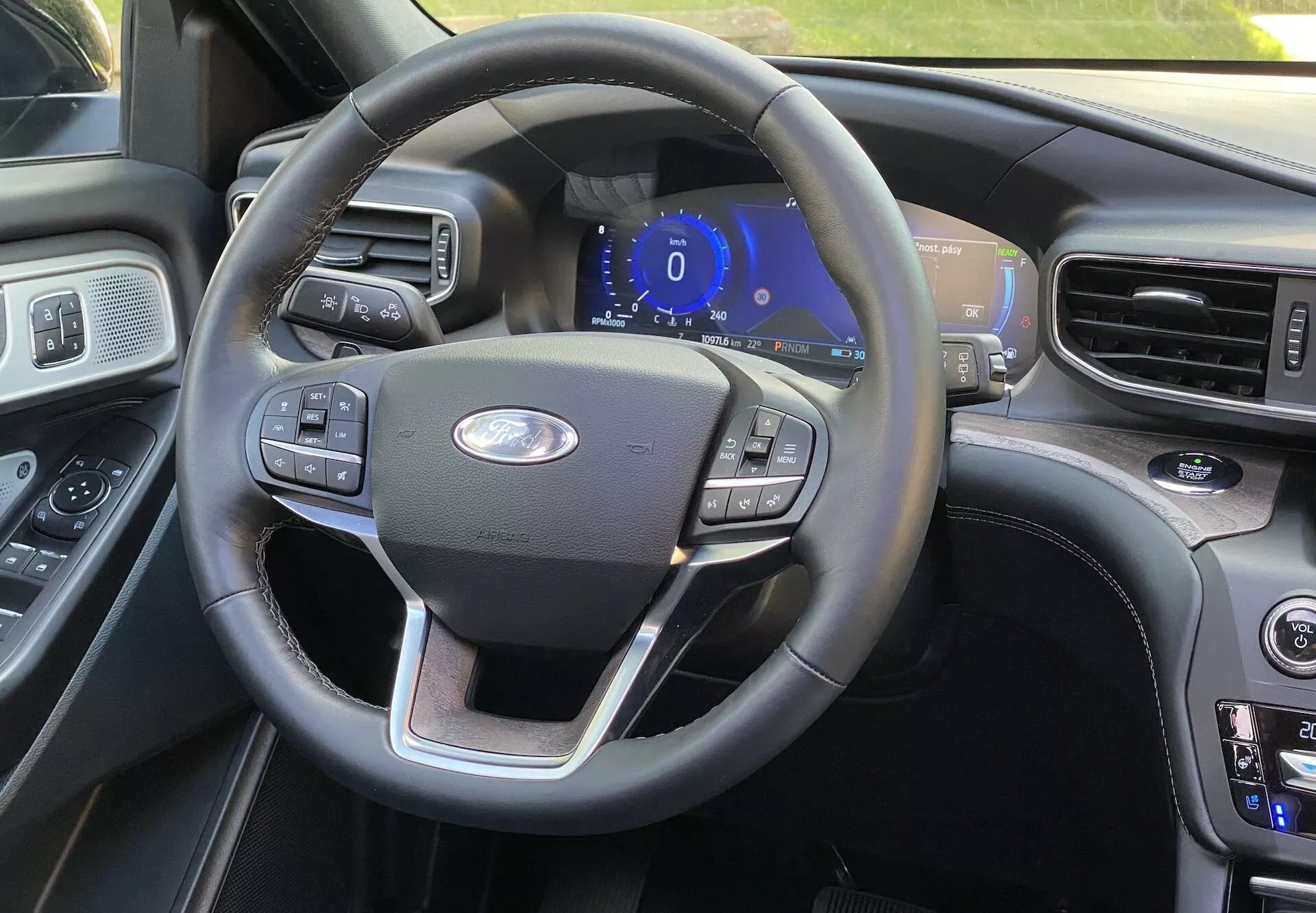 Pohľad na interiér Ford Explorer 2019 z pohľadu vodiča, ukazujúci trojramenný volant s multifunkčnými tlačidlami pre tempomat, nastavenia audia a pripojenie telefónu. V strede je logo Ford a modro osvetlený digitálny prístrojový panel naznačuje, že je vozidlo v režime príslušenstva alebo na štarte. Vpravo je viditeľná časť stredovej konzoly s tlačidlami na nastavenie klimatizácie a teploty, čo naznačuje duálnu klimatizáciu. Interiér obsahuje mix kože a dreveného obloženia pre prémiový pocit, s ďalším displejom ľahko viditeľným na okraji.
