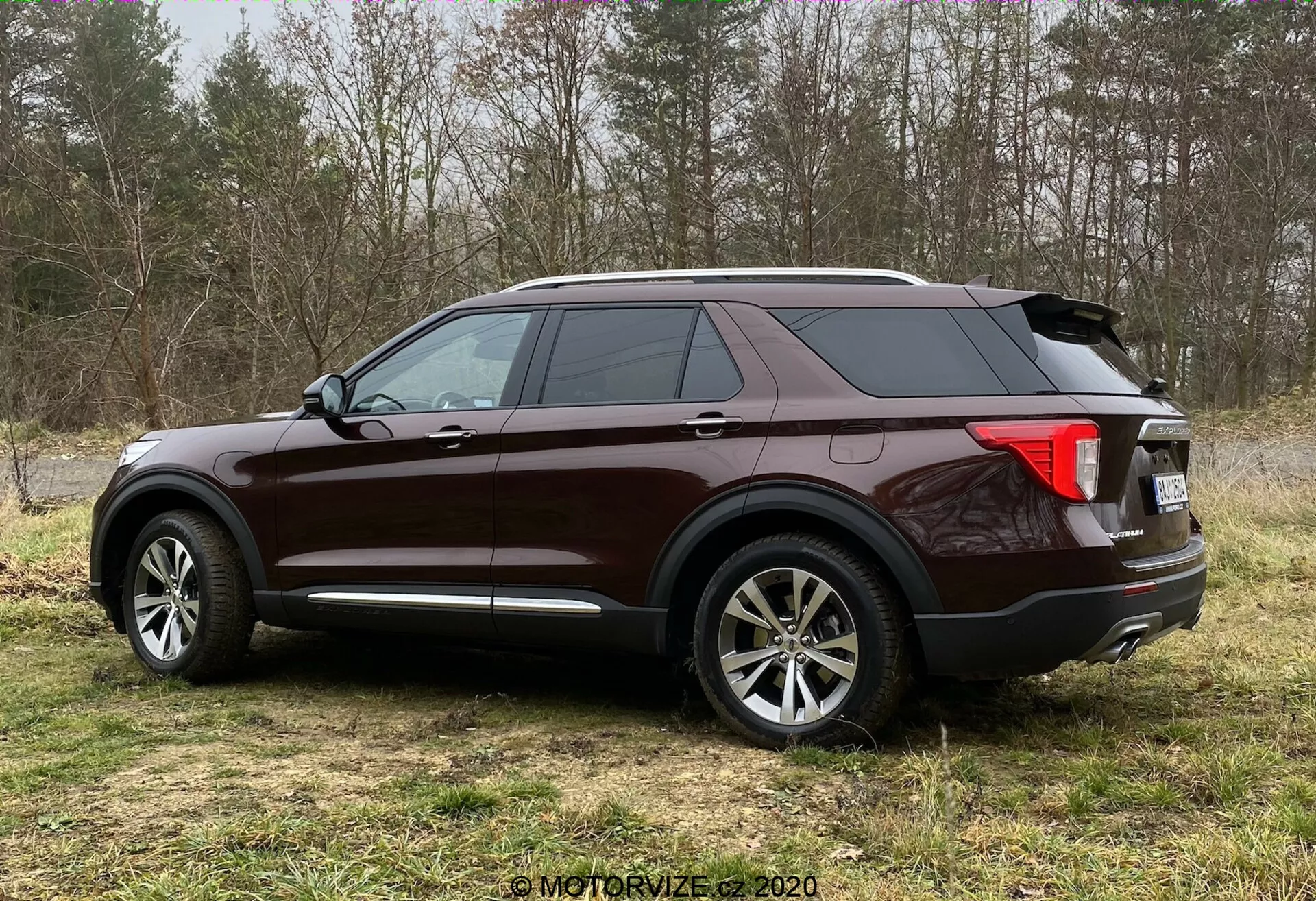 Bočný pohľad na SUV Ford Explorer 2019 v bohatom lesklom červenohnedom laku, stojaci na trávnatej ploche s pololesným pozadím. Trojštvrťový uhol ukazuje veľké viacramenné kolesá z ľahkej zliatiny, klesajúcu zadnú líniu strechy, tónované okná, výraznú bočnú charakterovú líniu, obopínajúce zadné svetlá pravdepodobne s LED, žraločiu anténu, chrómové lišty, strešné nosiče a dvojitý výfuk.