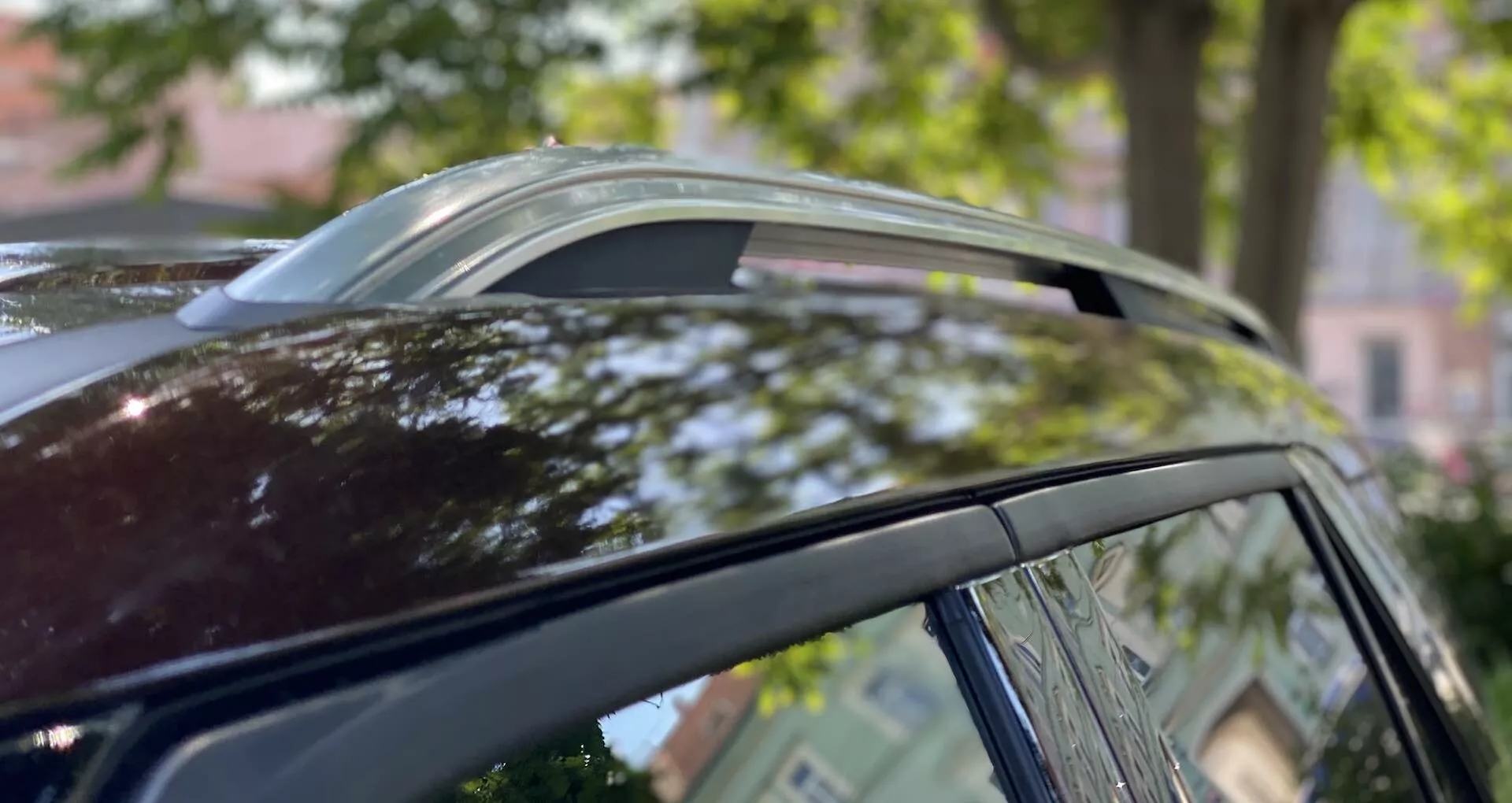 Blízky pohľad na strechu Ford Explorer 2019 z nízkeho uhla, zobrazujúci panoramatické strešné okno vo vetracej polohe, kovové okenné lišty a strešné nosiče pred rozmazaným pozadím stromov a architektúry.