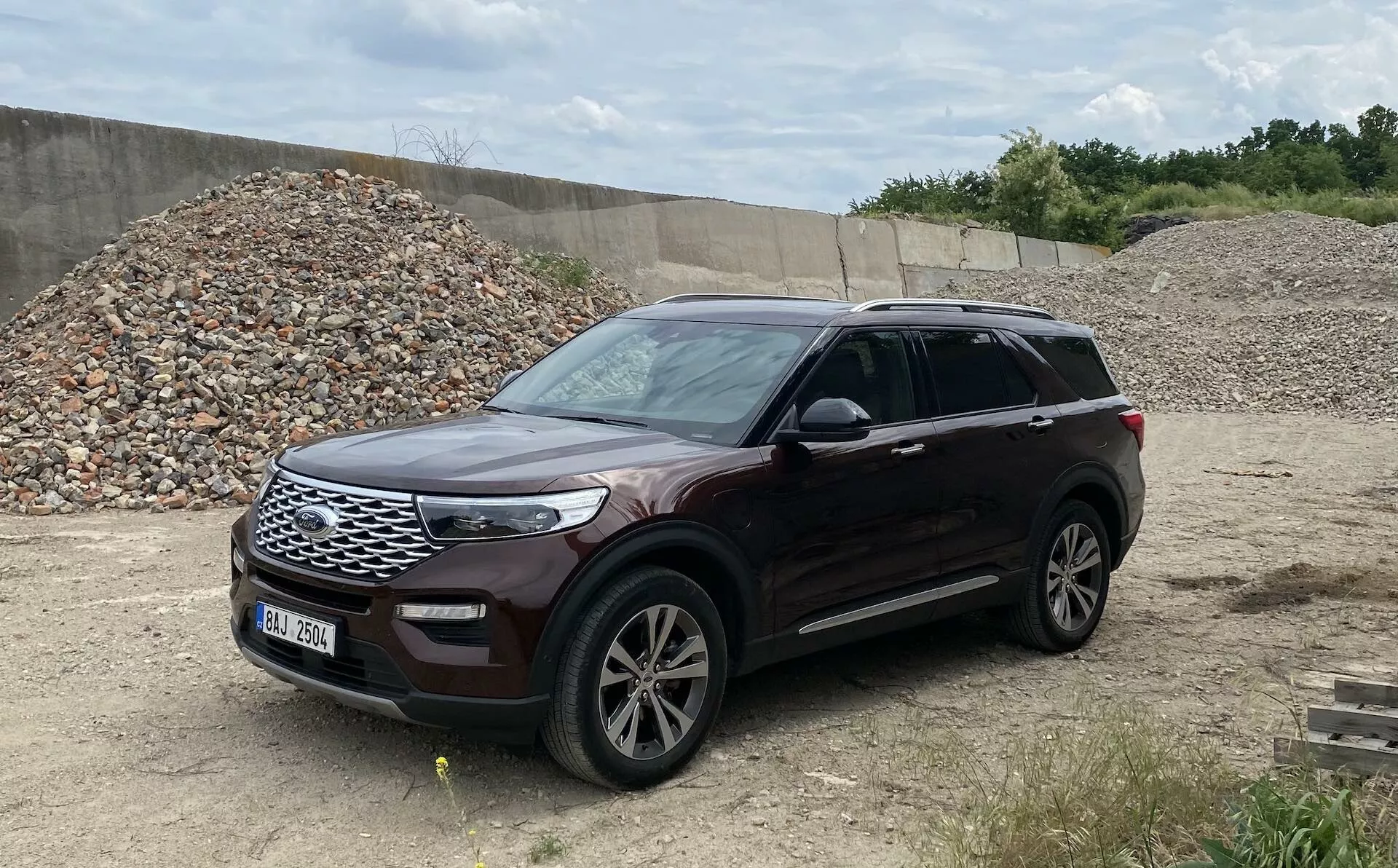 Obrázok zobrazuje Ford Explorer 2019, moderné SUV, zaparkované na nerovnom teréne pripomínajúcom stavenisko alebo off-road oblasť. Snímka z trojštvrťového pohľadu ukazuje prednú aj bočnú časť vozidla, vrátane mriežky chladiča, svetlometov, predného nárazníka, kapoty, zliatinových kolies, predných a zadných dverí, vonkajších spätných zrkadiel s integrovanými smerovými svetlami, blatníkov a okien. Vozidlo je orientované smerom doprava a metalický lak je jasne viditeľný, poskytuje komplexný pohľad na toto dobre udržiavané vozidlo s pozdĺžnymi nosníkmi na strechu, čo naznačuje možnosť prepravy ďalšieho nákladu.