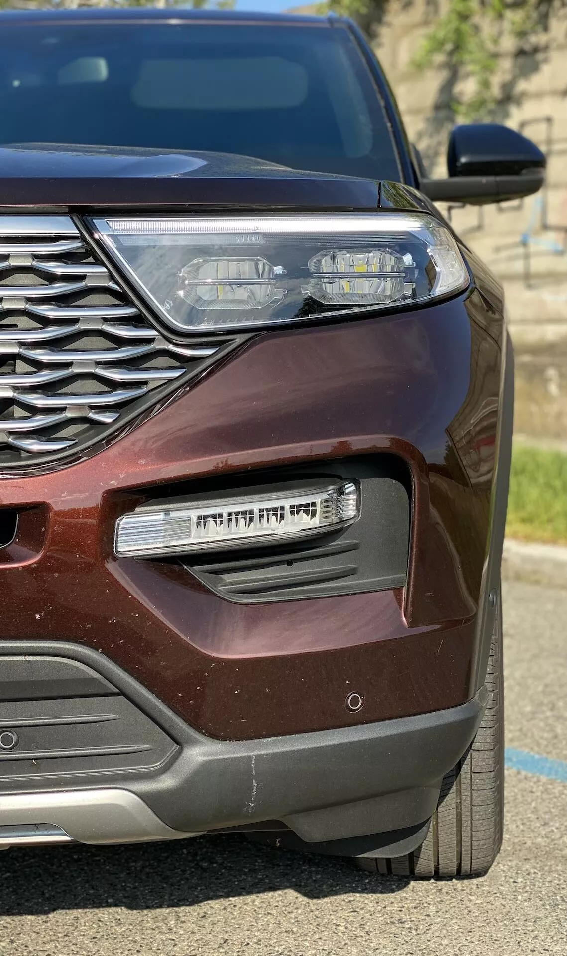 Detailný záber ľavej prednej štvrtiny moderného vozidla Ford Explorer 2019 v bordovej farbe, zobrazujúci zložitú chrómovú mriežku, elegantný LED svetlomet s dennými svetlami, chrómovaný kryt hmlového svetla a metalický lak, ktorý sa trblieta na slnku.