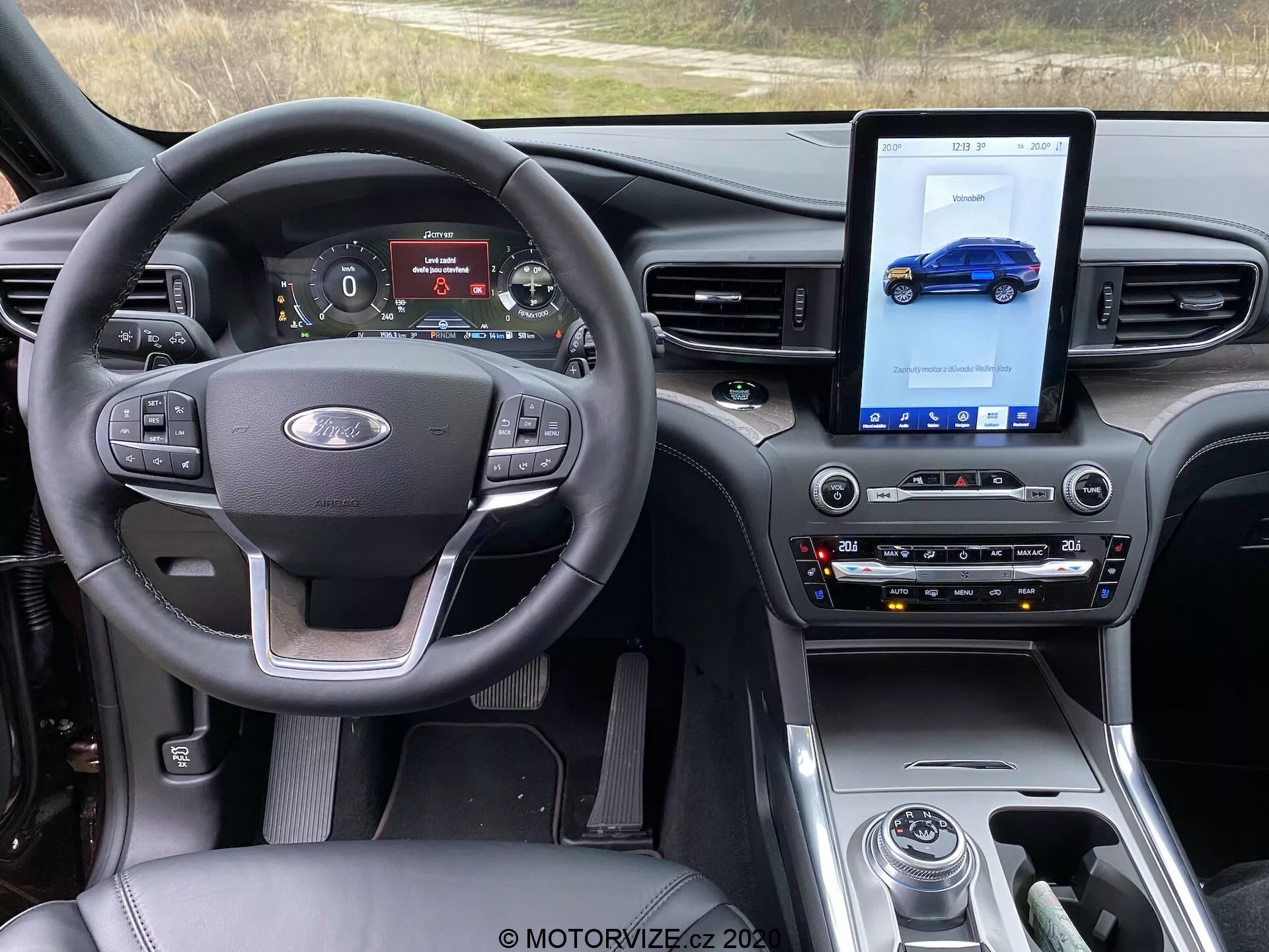 Interiér Fordu Explorer 2019 so zameraním na prednú kabínu z pohľadu vodiča. Obrázok zobrazuje volant s ovládacími tlačidlami na hlasitosť, hlasové príkazy a tempomat spolu s emblémom Ford v strede. Digitálny prístrojový panel zobrazuje rýchlomer, otáčkomer, režim jazdy a stav paliva. V stredovej konzole je veľký vertikálny dotykový displej infotainment systému, zobrazujúci modré vozidlo, tradičné ovládacie prvky na nastavenie klímy a audia a radiacu páku. Palubná doska má moderný dizajn s lesklými povrchmi, matnými prvkami a prvkami z brúseného kovu, ktoré zvyšujú moderný, prémiový dojem. Technologické prvky zahŕňajú multifunkčný volant, digitálny prístrojový panel, dotykový infotainment systém, automatickú klimatizáciu, volič jazdných režimov a ovládanie teploty sedadiel.
