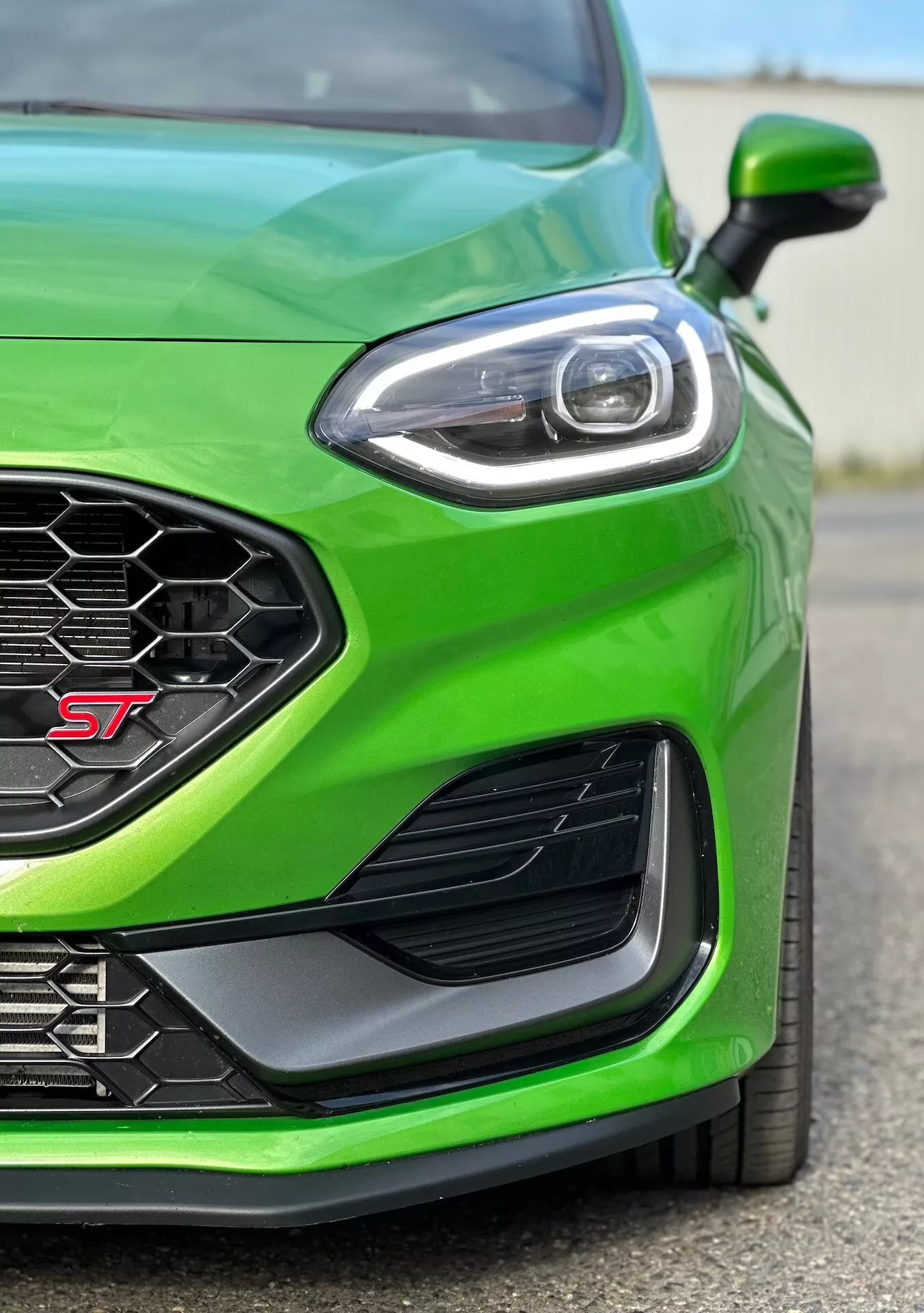 Detailný pohľad na zelený Ford Fiesta ST 2022 zameraný na výkonnostný predný ľavý štvrťrok, zdôrazňujúci športovo ladený vzhľad, medovcom vzorovanú mriežku pre optimálny prúd vzduchu, pokročilý dizajn LED svetlometov, aerodynamický bočný ventil a predný nárazník pre lepšie kanalizovanie vzduchu.