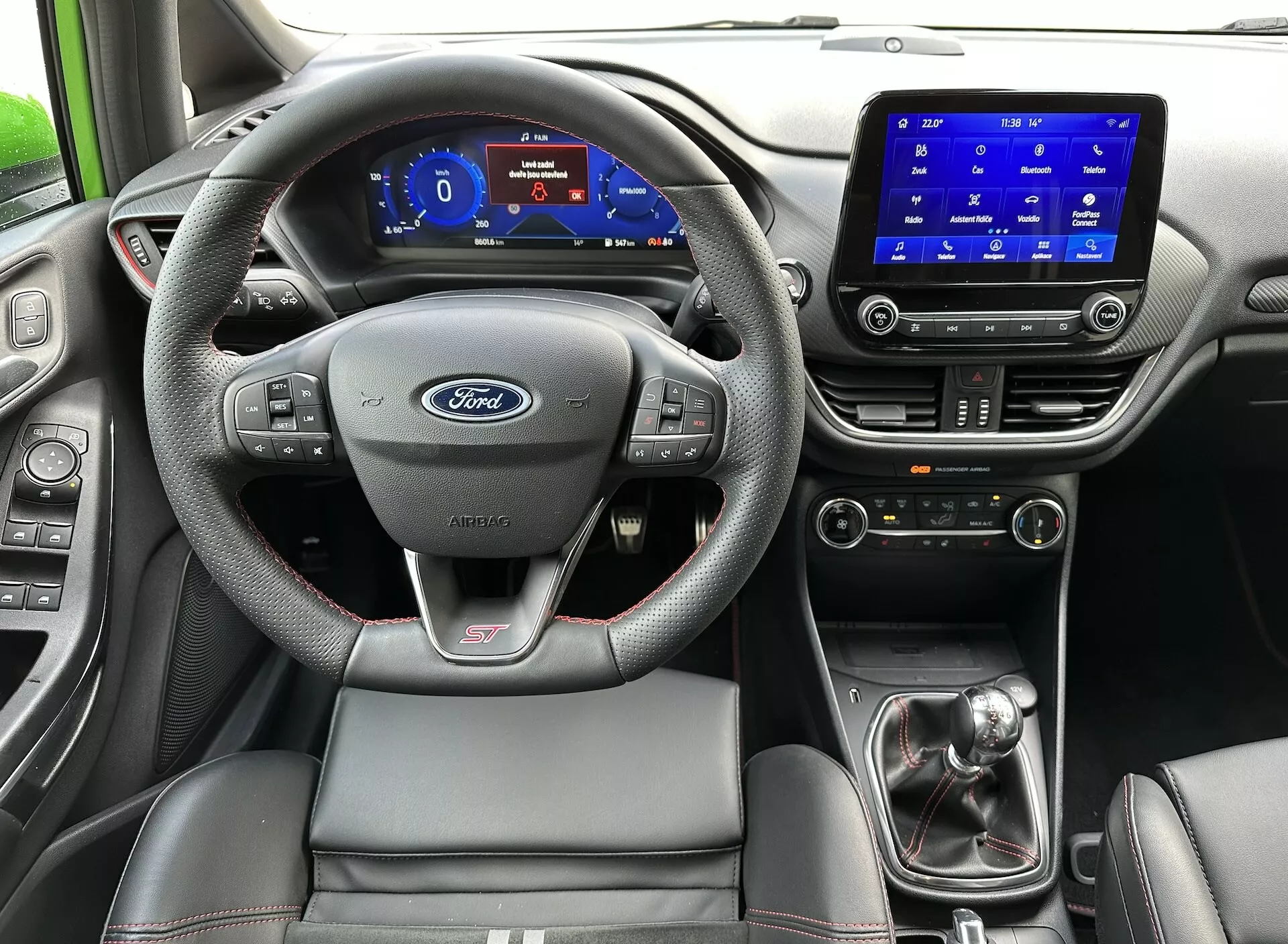 Interiér Ford Fiesta 2022 z pohľadu zadného sedadla, zdôrazňujúci volant s logom Ford a odznakom ST, digitálny prístrojový panel, dotykový systém infotainment, manuálnu radiacu páku a športové sedadlo vodiča s kontrastným prešívaním.