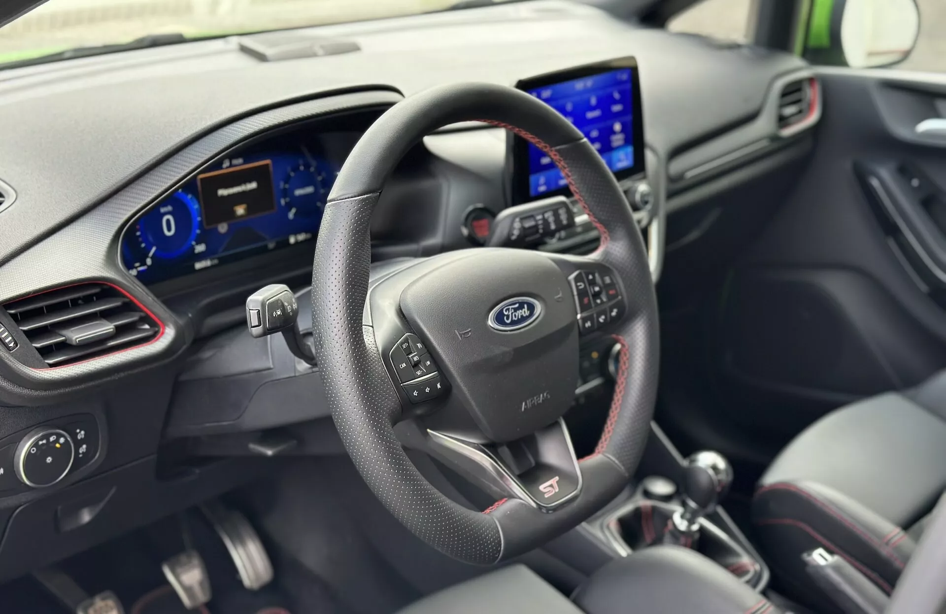 Interiér Ford Fiesta 2022 z pohľadu vodiča s koženým volantom s ovládacími tlačidlami, logom Ford, digitálnym prístrojovým panelom a moderným informačným systémom na palubnej doske