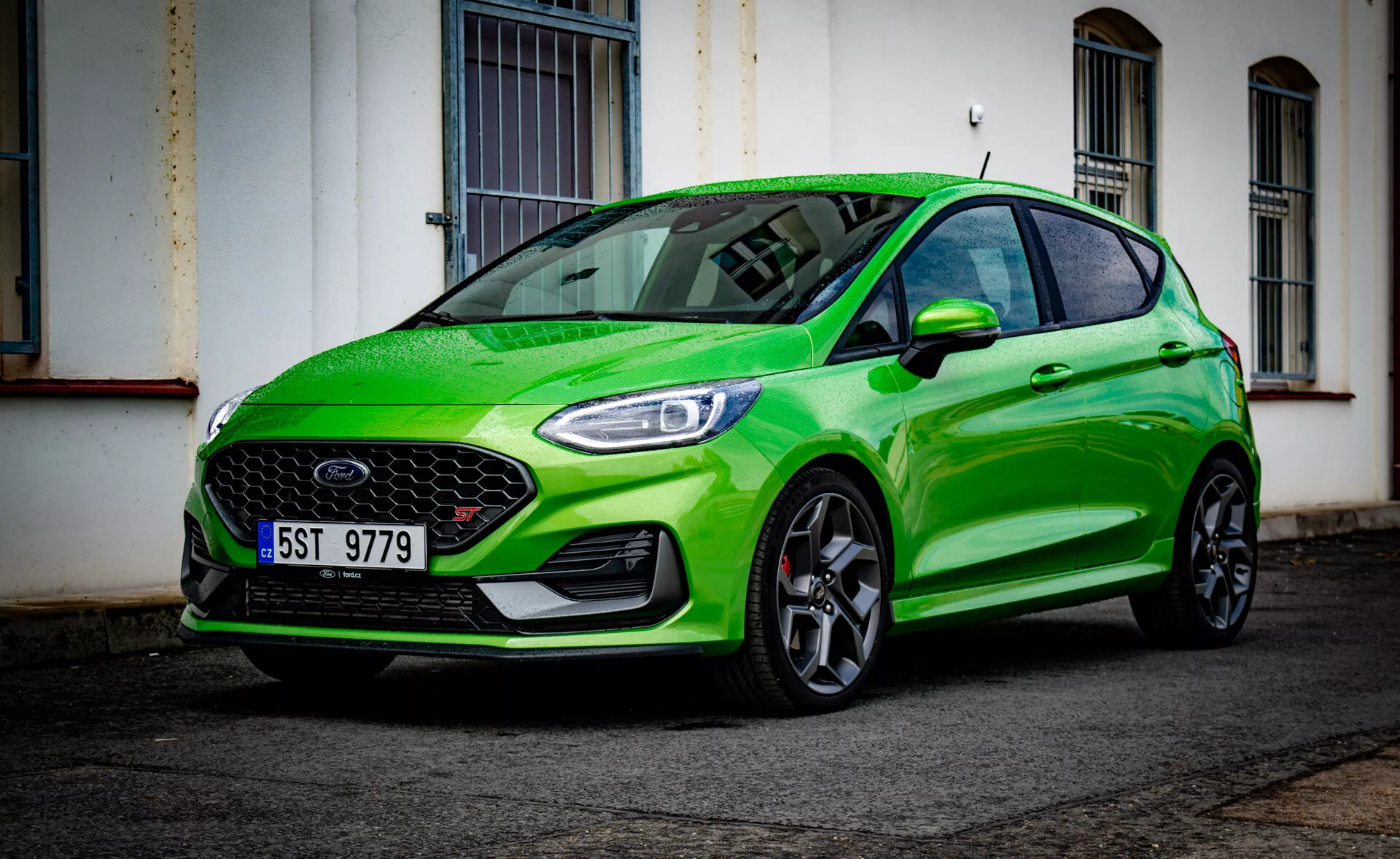 Ford Fiesta 2022 vo svetlozelenom prevedení, ktorá ukazuje svoj športový dizajn z nízkeho uhla pohľadu na prednú časť z troch štvrtín zdôrazňujúci predný nárazník, mriežku, svetlomety, kapotu, blatník, kolesá a odraz čelného skla na mestskom ulici po daždi