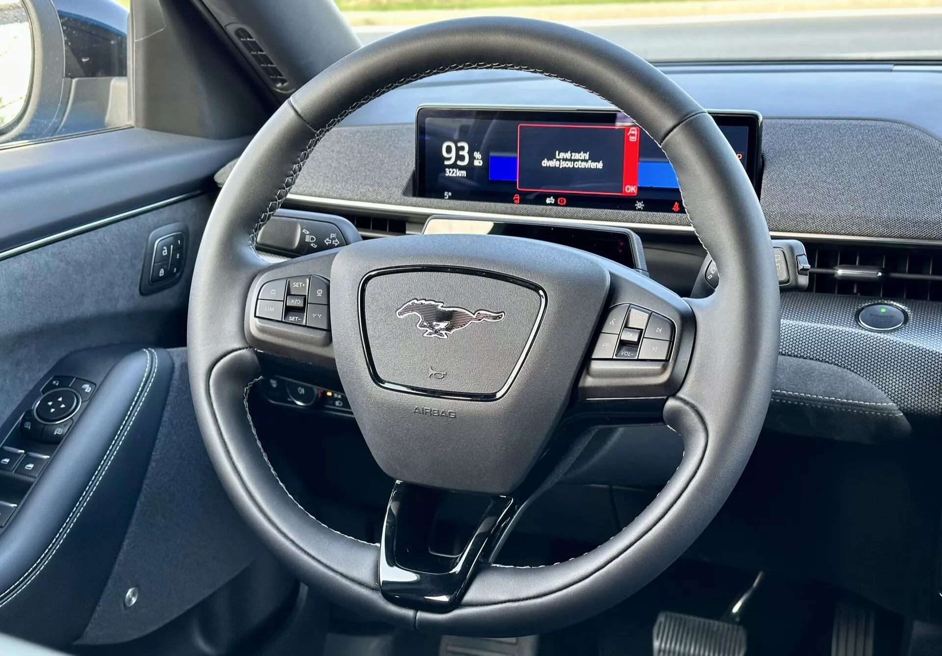 Interiér Ford Mustang Mach-E 2020 zvýrazňujúci moderný trojramenný volant s multifunkčnými tlačidlami a digitálny prístrojový panel z pohľadu vodiča, ukazuje ovládacie prvky, emblém a náznaky navigácie.