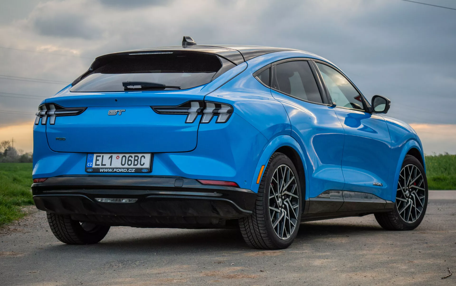 Modré elektrické SUV Ford Mustang Mach-E 2020 zobrazené z trojštvrťovej zadnej perspektívy, zdôrazňujúce jeho štýlový aerodynamický dizajn, moderné zadné LED svetlá, zliatinové kolesá s viacerými špicami, označenie elektrického vozidla a technologické funkcie