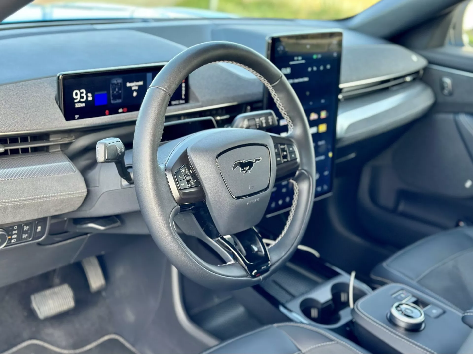 Interiérový pohľad na Ford Mustang Mach-E 2020 zameraný na kokpit vodiča z pohľadu spolujazdca, predstavujúci volant s emblémom Mustang, digitálny palubný displej v režime Park zobrazujúci dojazd 93 míľ, a vertikálne orientovaný centrálny dotykový displej integrovaný s pokročilými funkciami infotainmentu.