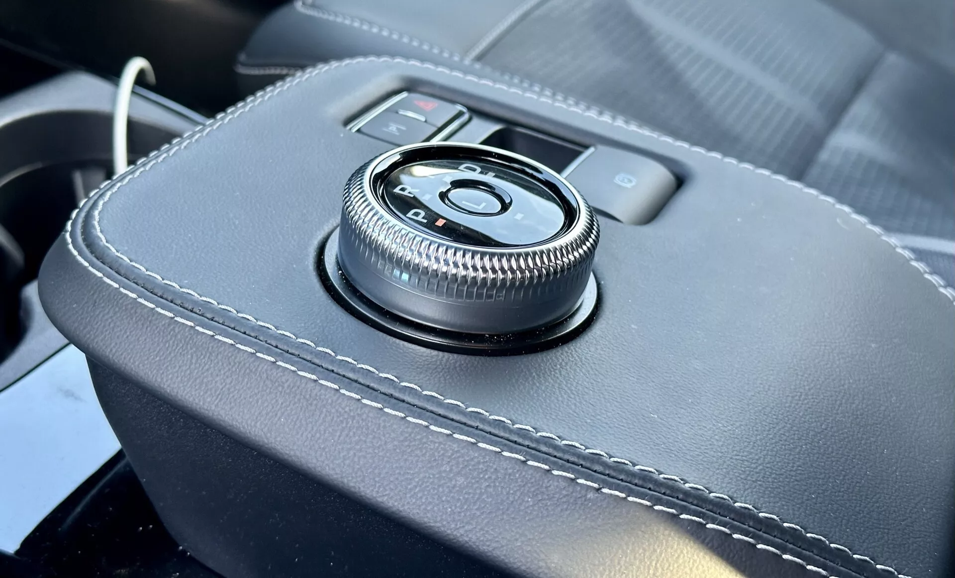 Detailný pohľad na interiér Ford Mustang Mach-E 2020 s otočným ovládačom na stredovej konzole na povrchu obalenom kožou s bielym prešívaním, poskytujúci ergonomické rozhranie pre informačno-zábavný systém