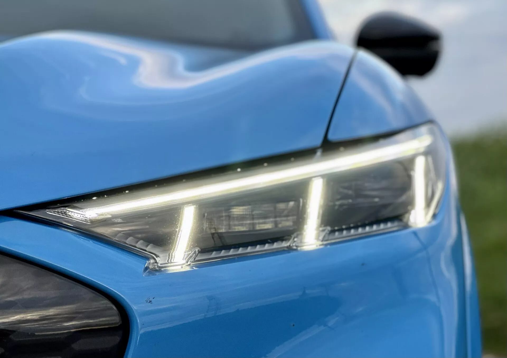 Detailný pohľad na prednú ľavú časť Ford Mustang-Mach-E 2020 s dôrazom na elegantné LED svetlomety, živú modrú farbu, dynamické línie karosérie a časť predného nárazníka a bočného zrkadla, čo zvýrazňuje moderný dizajn a estetiku.