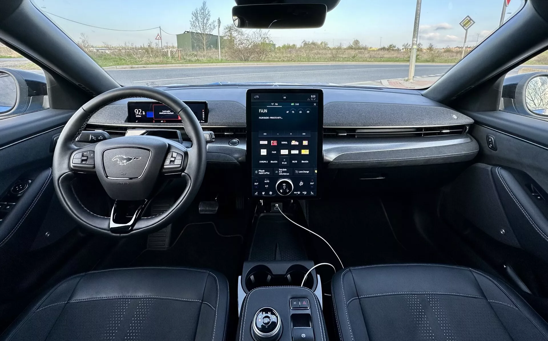 Pohľad z prvej osoby na interiér Ford Mustang Mach-E 2020, ukazujúci volant na ľavej strane s plochým dnom a emblémom, digitálny prístrojový panel, veľký vertikálny dotykový infotainment systém na stredovej konzole s ovládacími gombíkmi klimatizácie nižšie, USB porty a luxusné čalúnenie v tmavých tónoch s detailným šitím, naznačujúce pokročilé bezpečnostné a asistenčné funkcie, s viditeľným vidieckym pozadím cez čelné sklo.