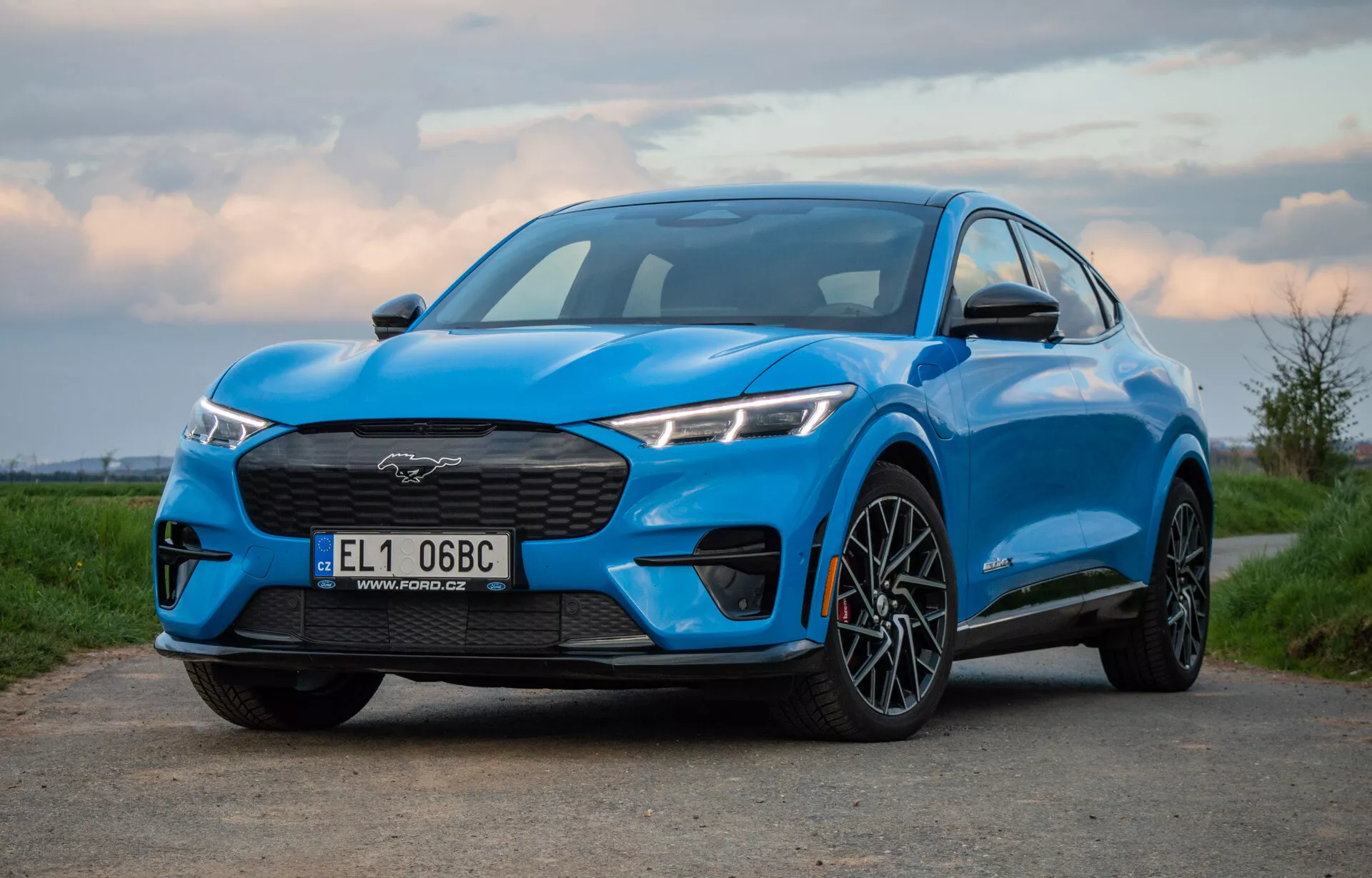 Modrý Ford Mustang Mach-E 2020 odfotografovaný z nízkeho a mierne z boku uhla, zobrazujúci mriežku, svetlomety, kapotu a profil vozidla vrátane predného kolesa a blatníka, zdôrazňujúce jeho výkon a aerodynamický dizajn na úsvite alebo súmraku.