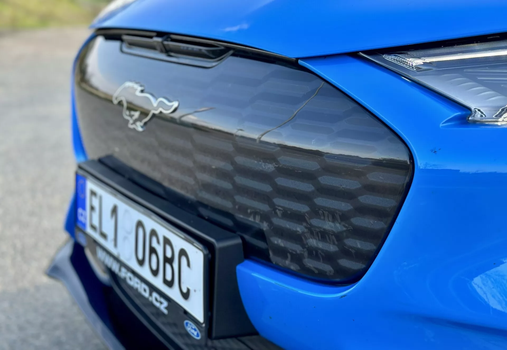 Detailný záber na zadnú časť modrého športového auta Ford Mustang Mach-E 2020 ukazujúci chrómový emblém koňa, spoiler z uhlíkových vlákien, integrovanú níšu pre poznávaciu značku a čiastočný pohľad na LED zadné svetlá, zdôrazňujúci aerodynamické a výkonnostné vlastnosti auta.
