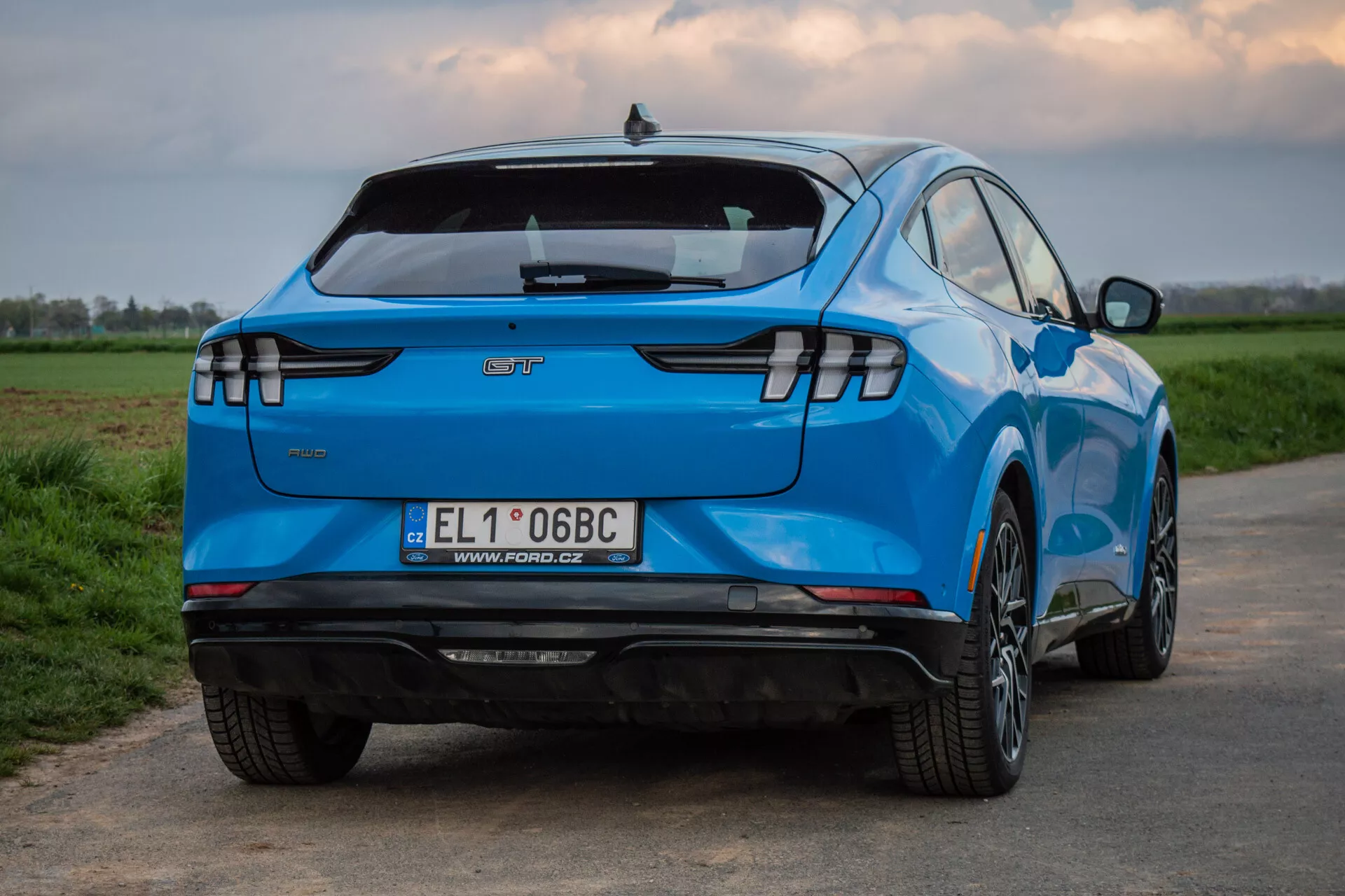 Ford Mustang-Mach-E 2020 nafotený zozadu a z trojštvrťového uhla, zobrazujúc jeho charakteristický dizajn, vrátane horizontálnych LED zadných svetiel, emblému výrobcu, integrovaného zadného spojlera, zadného nárazníka vybaveného senzormi bez viditeľného výfuku naznačujúceho elektrické vozidlo, európskej poznávacej značky, viacprvkových zliatinových kolies, žiarivomodrej farby a aerodynamickej kontúrovanej karosérie na pozadí vidieckej krajiny.