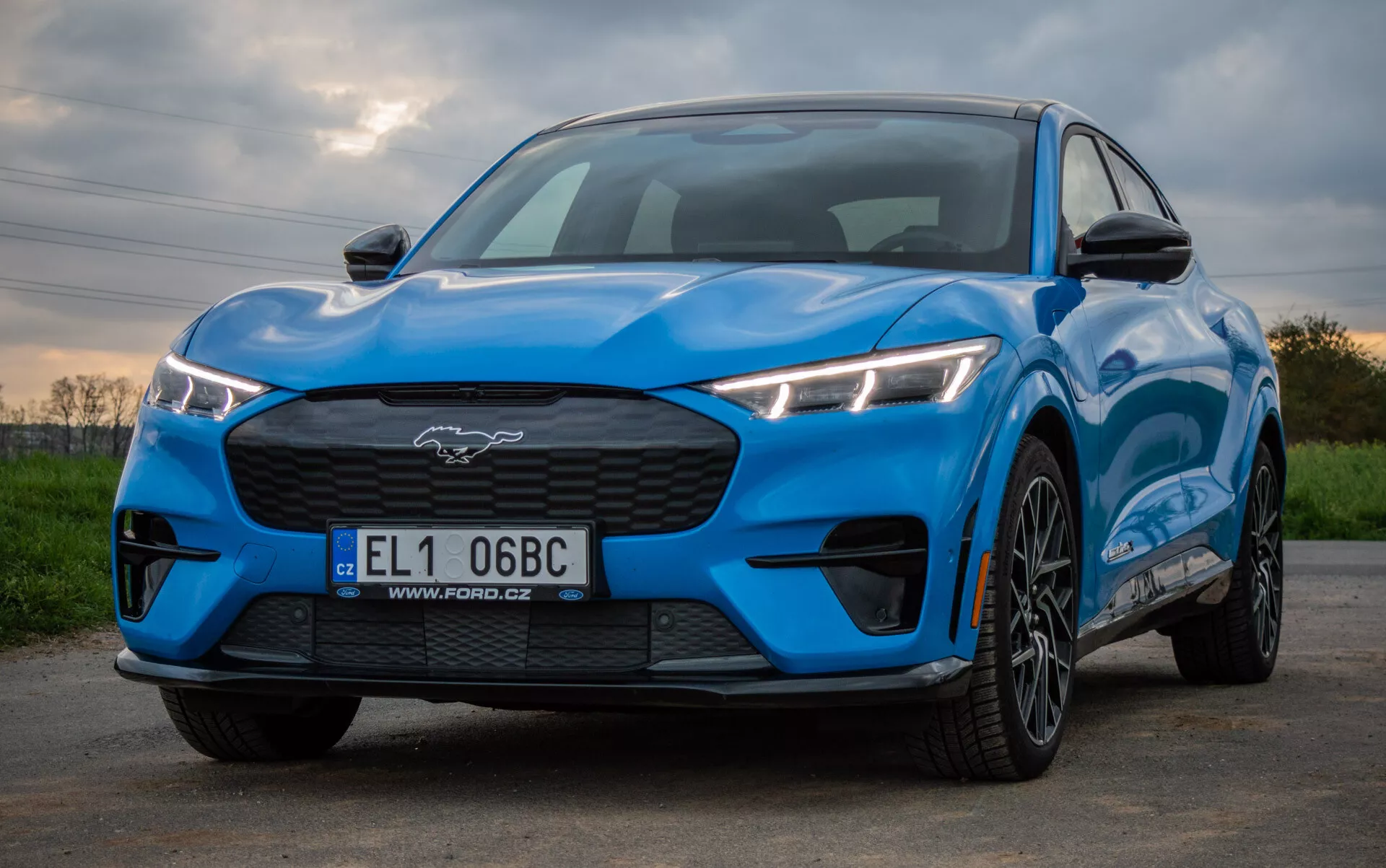 Ford Mustang Mach-E rok 2020 v jasne modrej farbe predvádzajúci športový a aerodynamický dizajn z nízkeho uhla pohľadu spredu a zboku, zvýrazňujúci uhlové LED svetlomety, zatvorenú mriežku, profilovanú kapotu a kolesá z ľahkej zliatiny s viacramenným dizajnom proti prírodnému pozadiu.