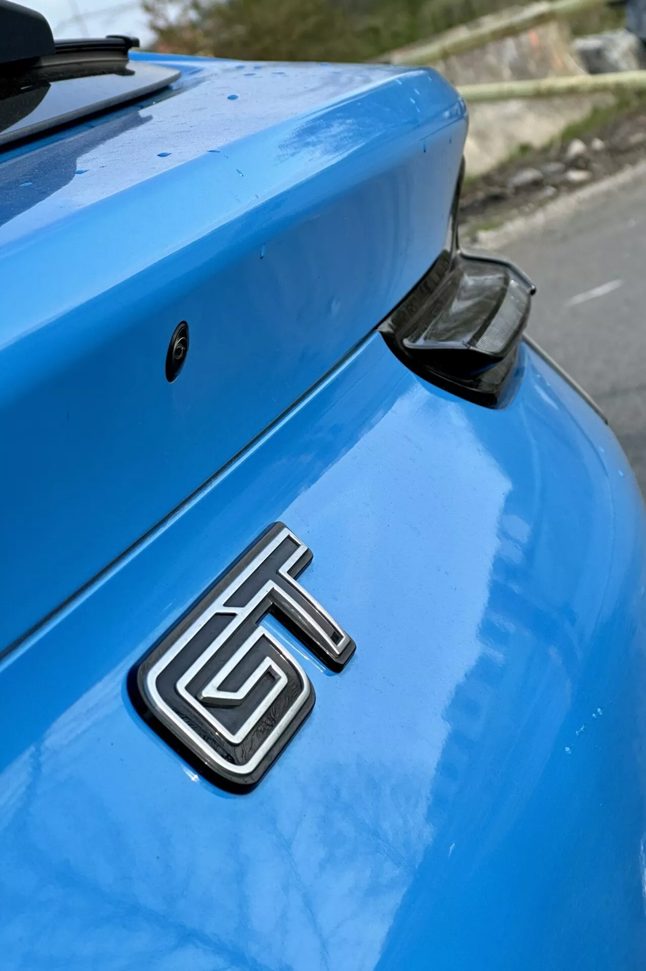 Detailný záber na zadnú časť modrého Ford Mustang Mach-E z roku 2020 so zameraním na kovovú značku GT, zakrivený kufor a aerodynamický zadný spojler s čiernym koncovým krytom na jemne rozostrenom pozadí.
