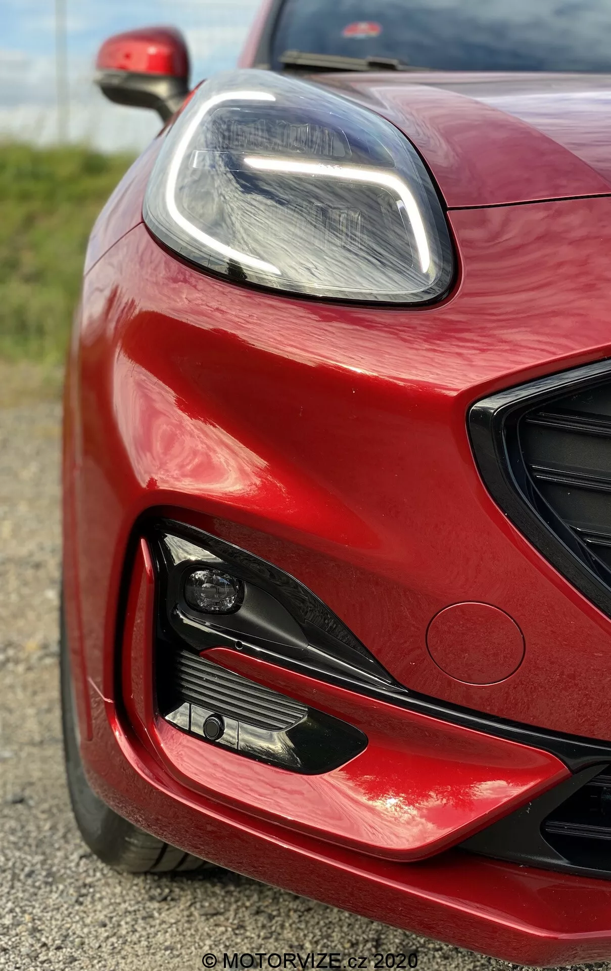 Detailný záber na prednú ľavú časť vozidla Ford Puma 2020 v žiarivej červenej farbe s lesklým povrchom, zobrazujúci elegantný LED svetlomet, veľké nasávacie otvory a hmlové svetlo, zdôrazňujúci jeho moderný, výkonnostne orientovaný dizajn.
